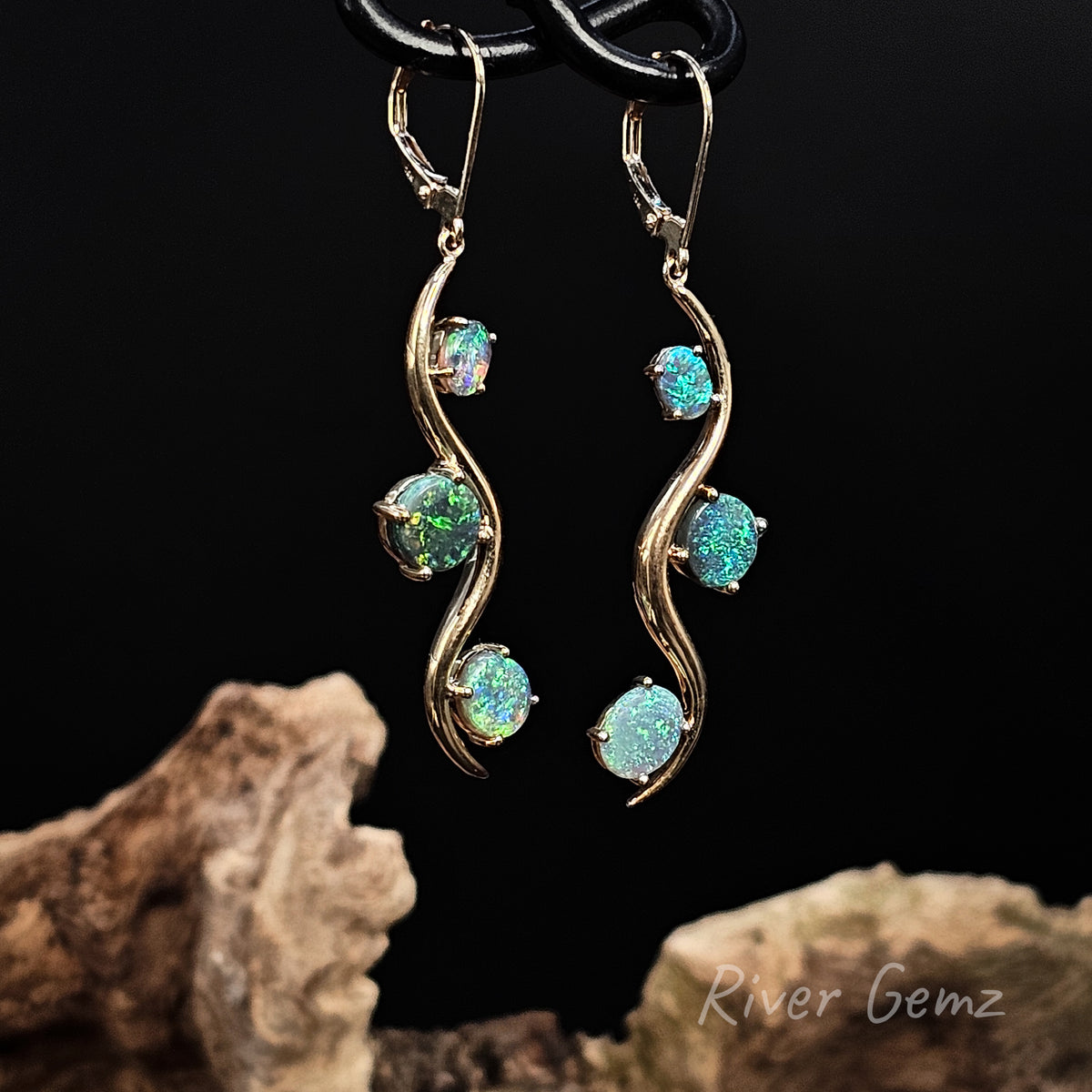3 opals on shepherd hook gold earrings against a black background with a light coloured piece of drift wood at the base.