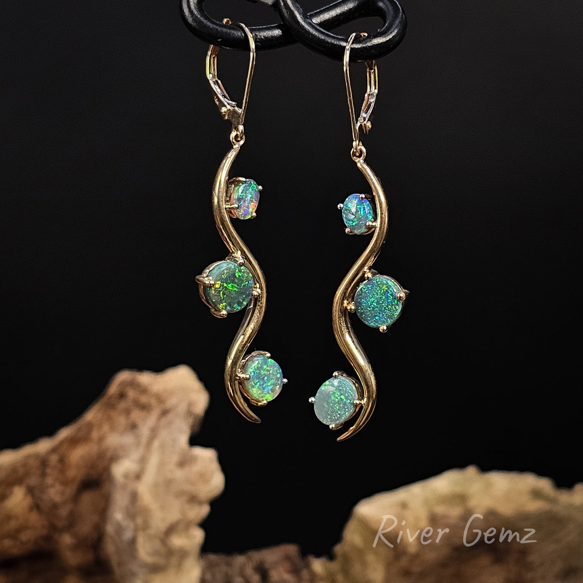 Three oval shaped blue-green dark opals claw set each side of a curve design pair of dangly yellow gold earrings against a black background.
