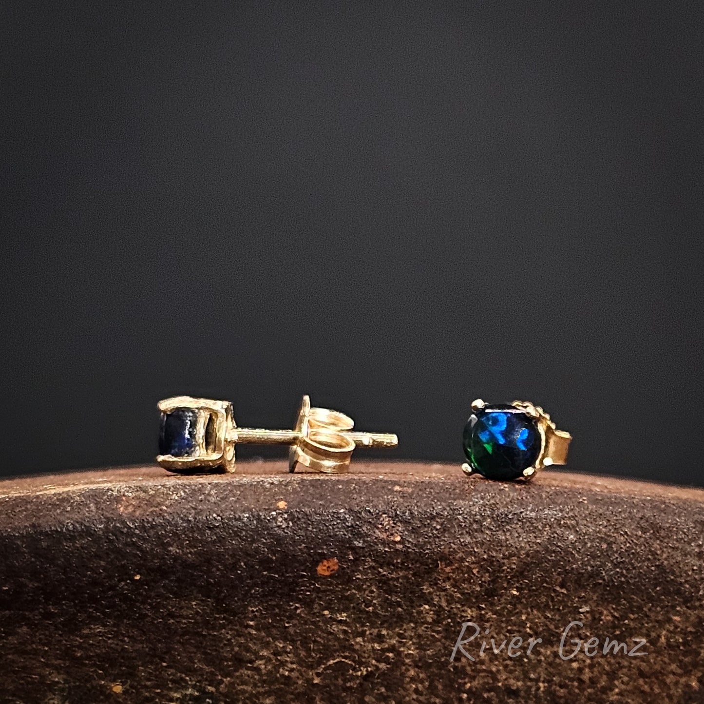 Gold stud earrings with black opals on a dark background, featuring the brand 'River Gemz'.