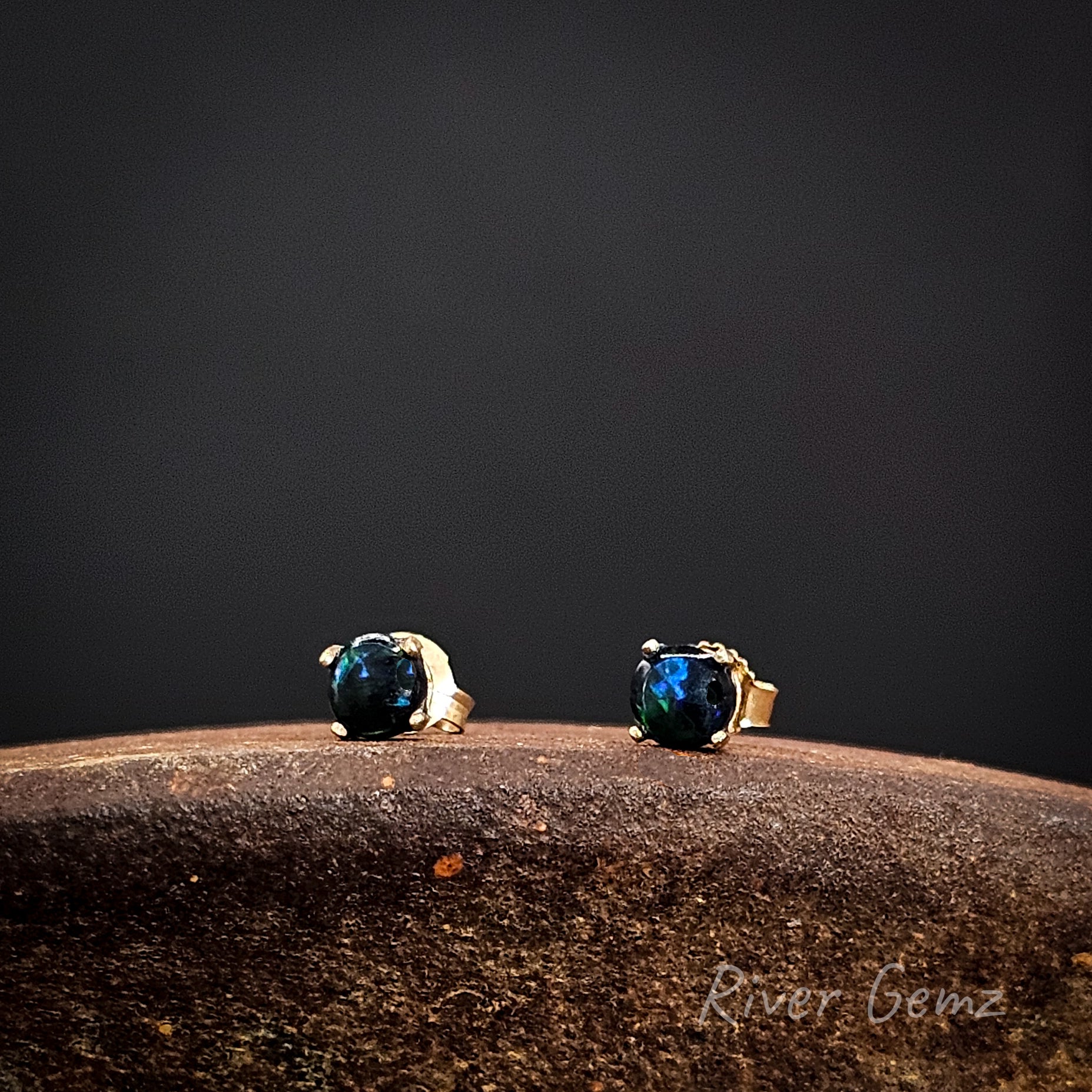 Pair of gold earrings with round dark opals on a dark background, featuring the brand 'River Gemz'.