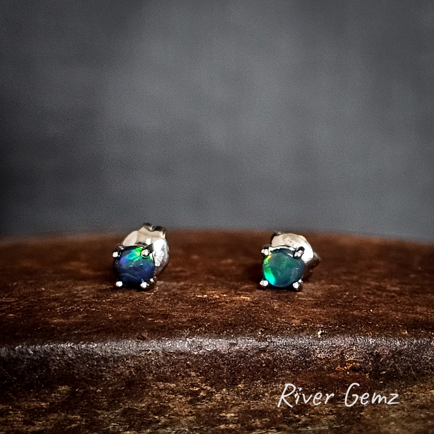 Opal stud earrings on a cast iron surface with a dark background, featuring the brand 'River Gemz'.