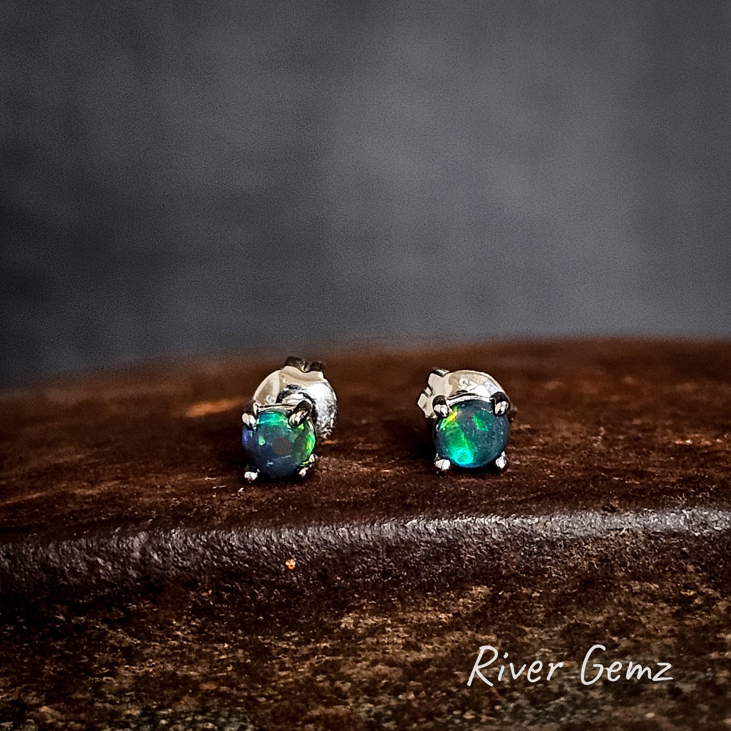 Pair of sterling silver opal stud earrings on a cast iron surface with 'River Gemz' branding.