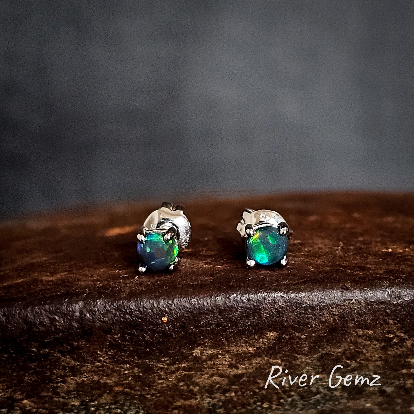 Pair of sterling silver opal stud earrings on a cast iron surface with 'River Gemz' branding.