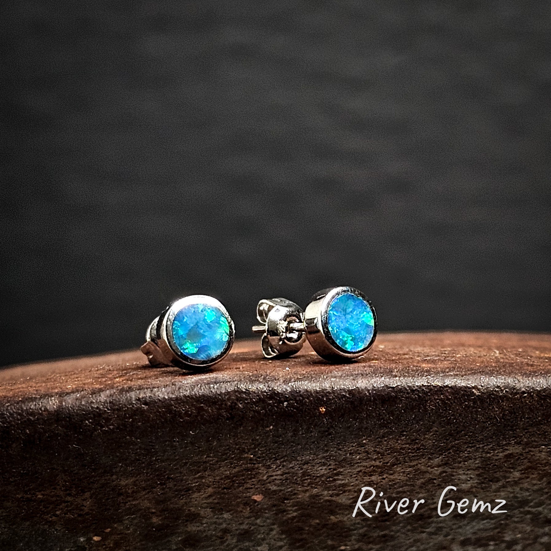 Pair of blue black opal stud earrings on a dark surface with 'River Gemz' branding.