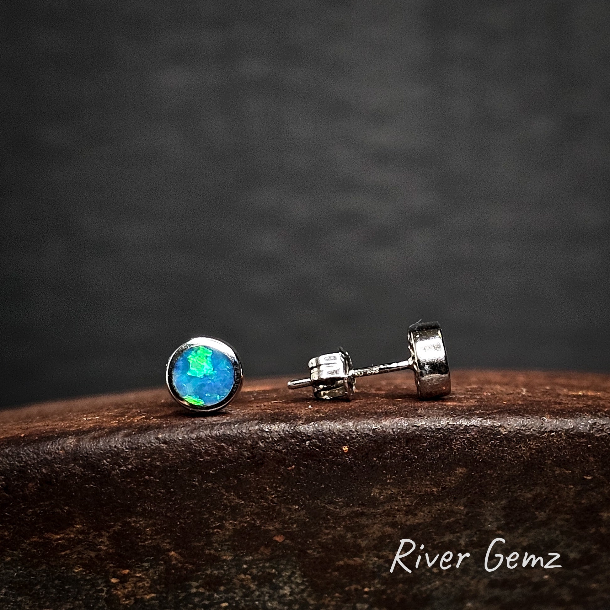 Blue and green round opals besel set in sterling silver stud earrings on a dark surface with 'River Gemz' branding.