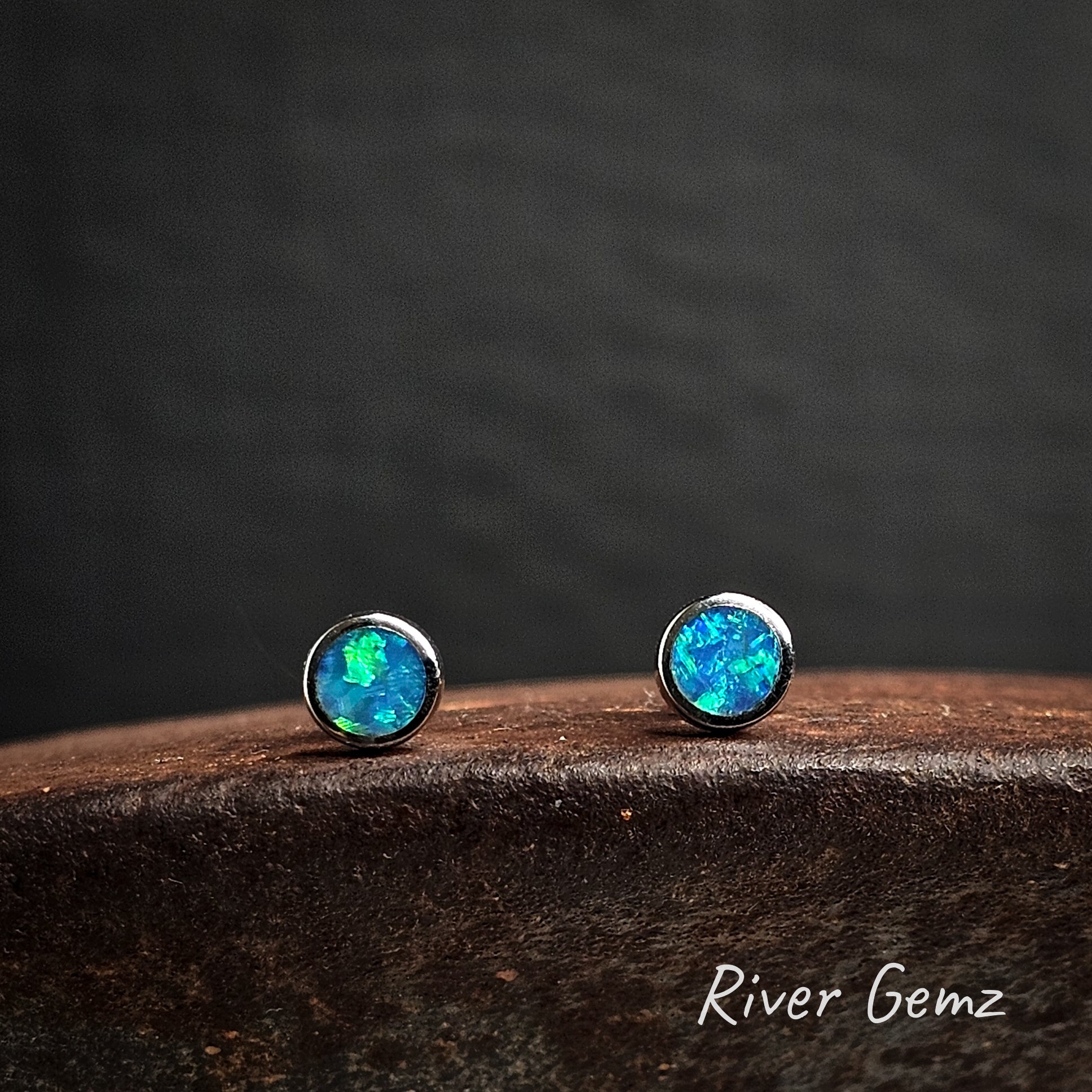 Opal stud earrings set in silver on a cast iron surface with 'River Gemz' branding.