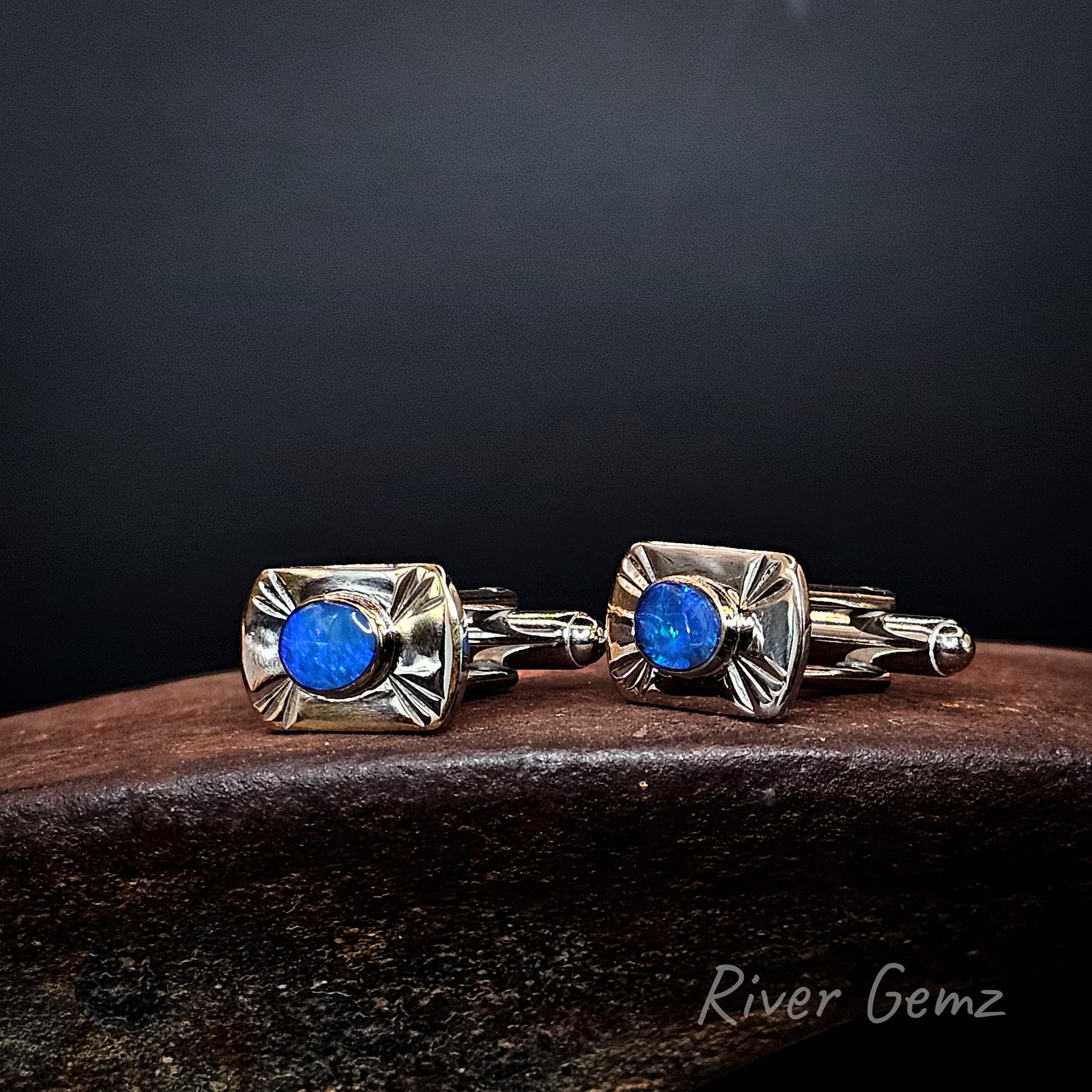 A pair of men's cufflinks by River Gemz, featuring square silver-toned metal settings with a textured sunburst pattern and a round brilliant-cut blue opal gemstone in the center, resting on a dark wooden surface against a dark background.