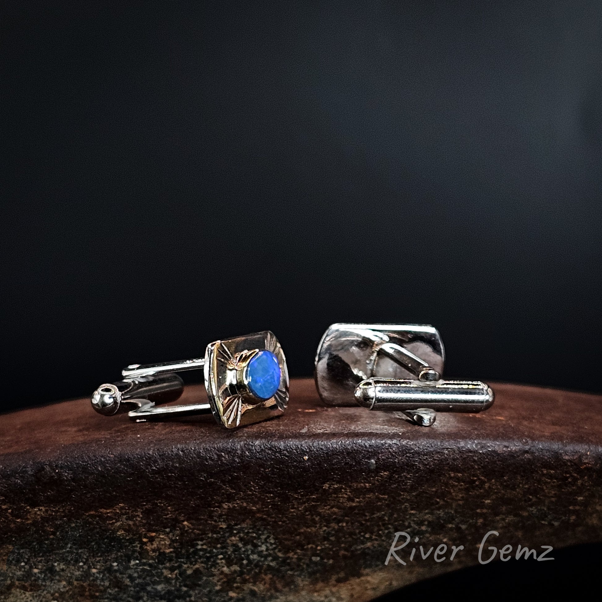 A pair of men's cufflinks displayed on a dark wooden surface against a black background. One cufflink is shown from the front, featuring an oval bezel-set blue dark opal with a sunburst pattern around it, attached to a silver-toned bar.