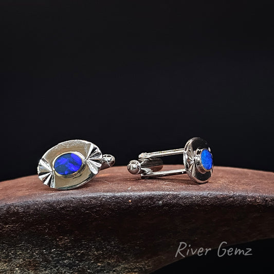 A pair of silver cufflinks with oval blue opal centers, displayed on a dark wooden surface against a black background. The brand name 'River Gemz' is visible as a watermark in the bottom right corner.