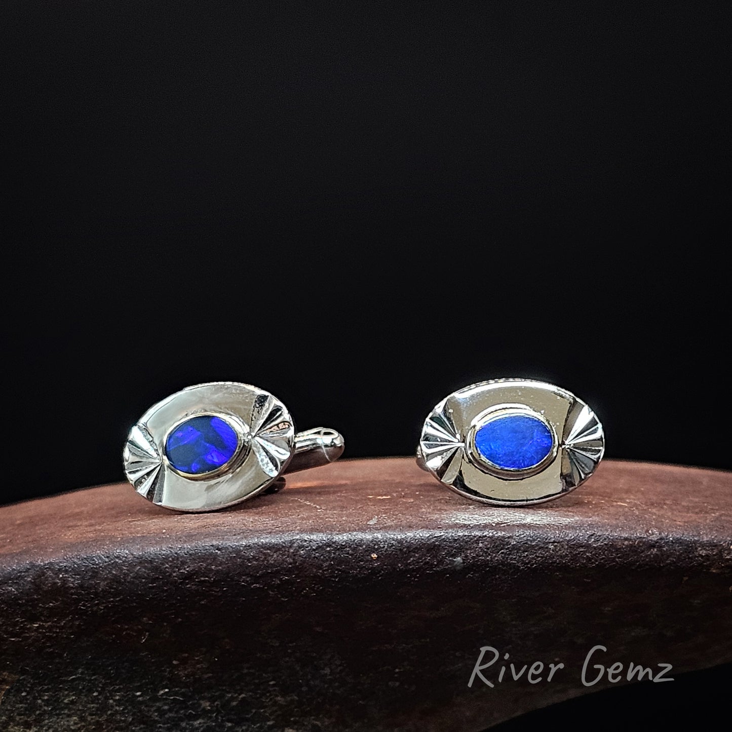 A pair of oval silver stud cufflinks with vibrant blue gemstone centers, displayed on a dark wooden block against a solid black background. The brand name 'River Gemz' is visible in the bottom right corner.