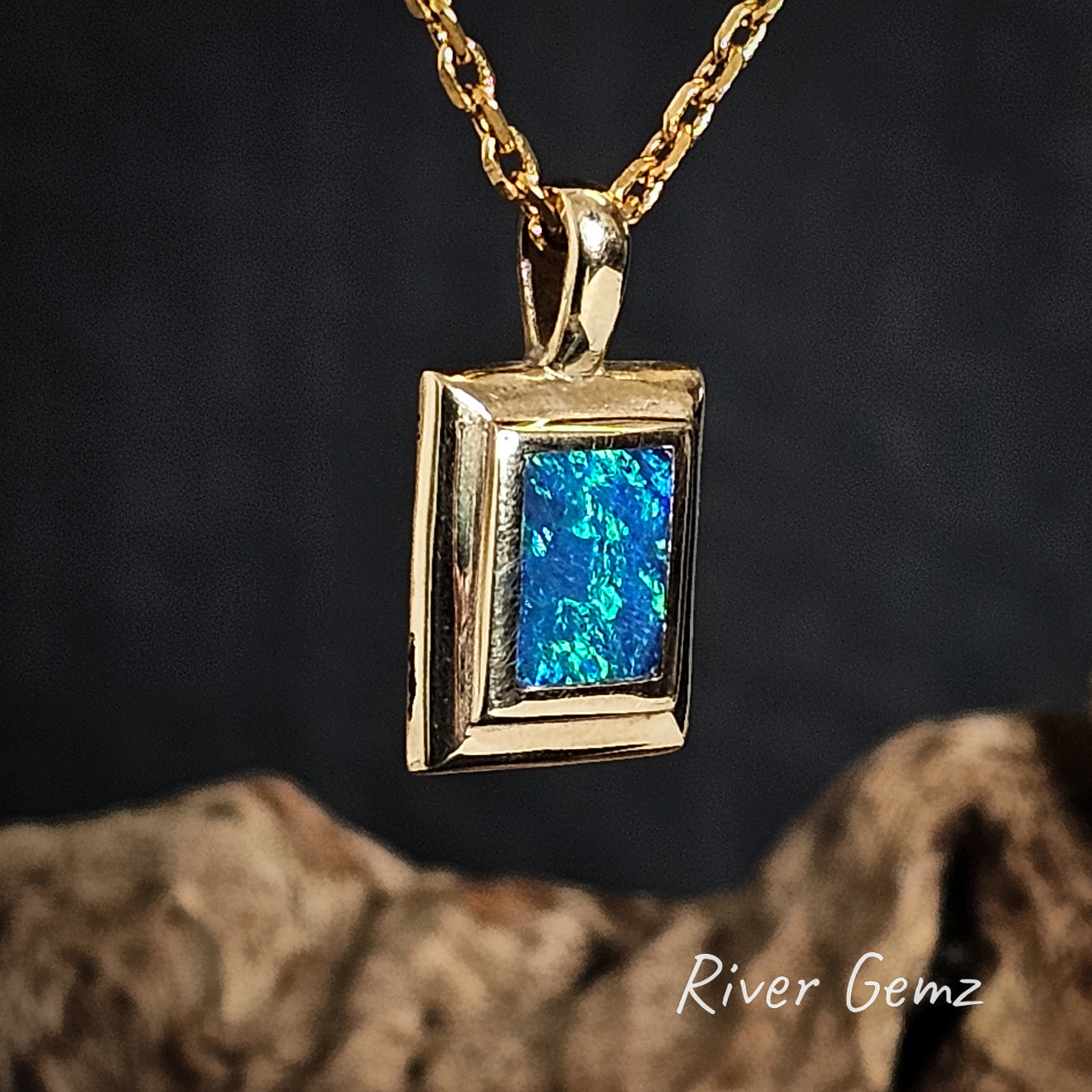 Gold pendant with a rectangular blue and green crystal opal on a dark background, featuring 'River Gemz' branding.