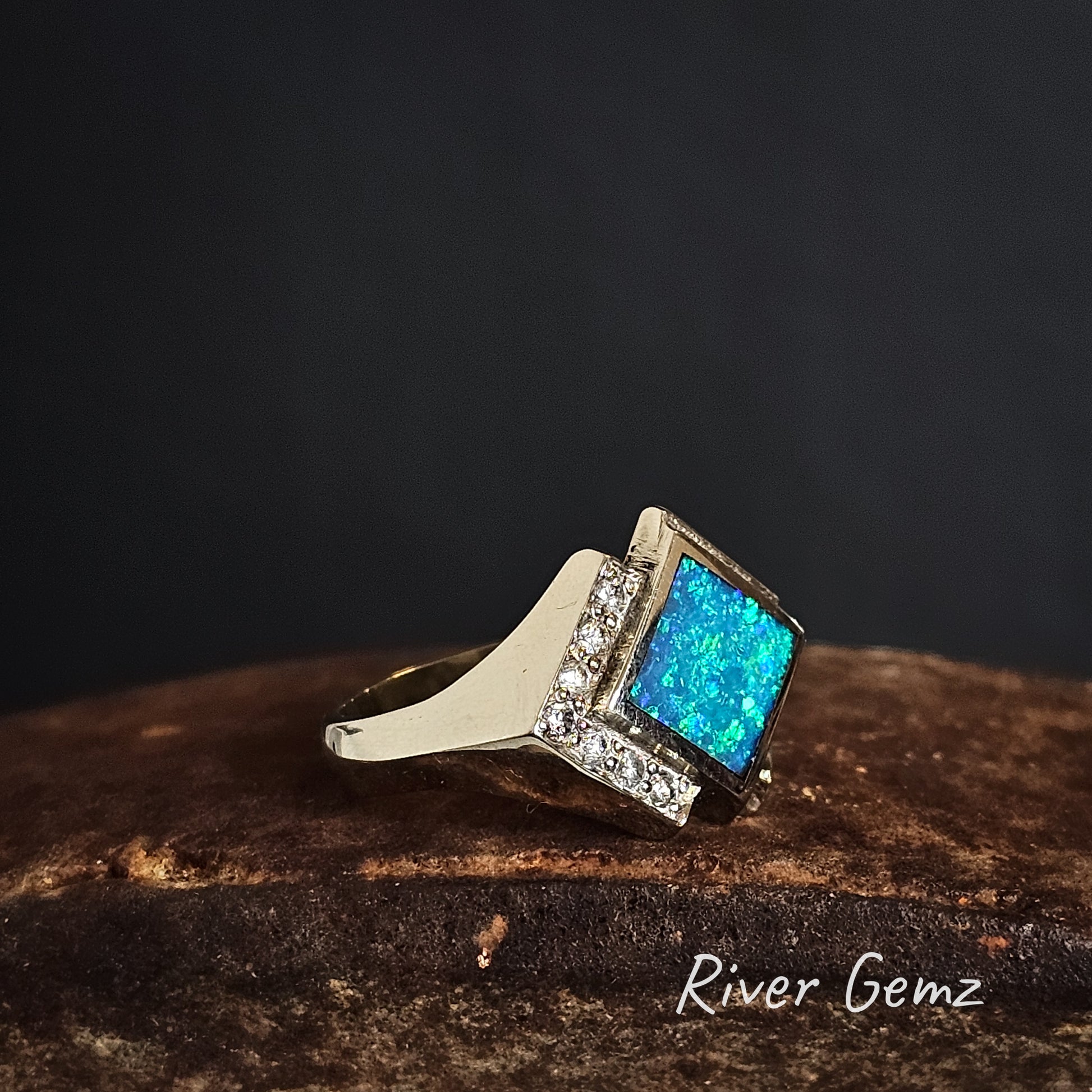 Ring with blue gemstone and diamonds on a dark background, branded 'River Gemz'.
