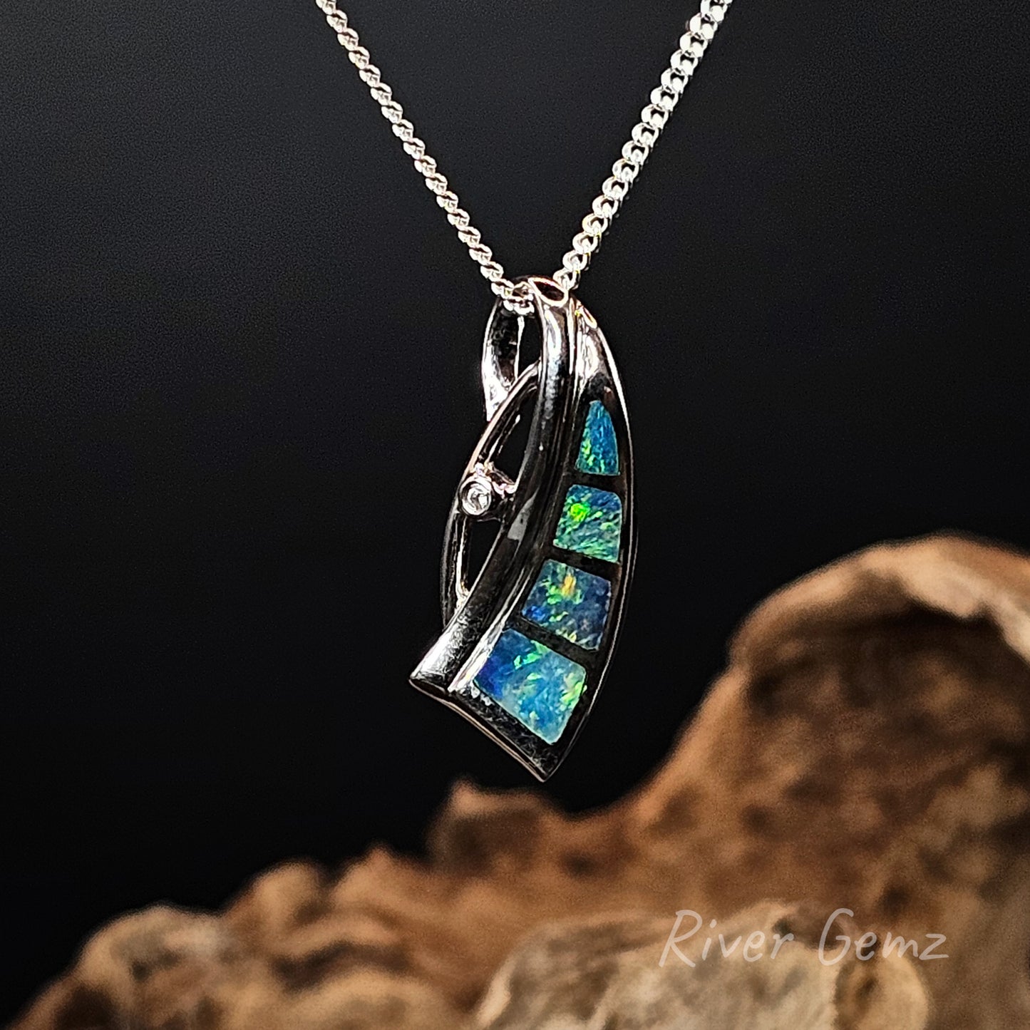 Silver pendant with channel set blue and green light opals and a white topaz highlight on a chain against a dark background