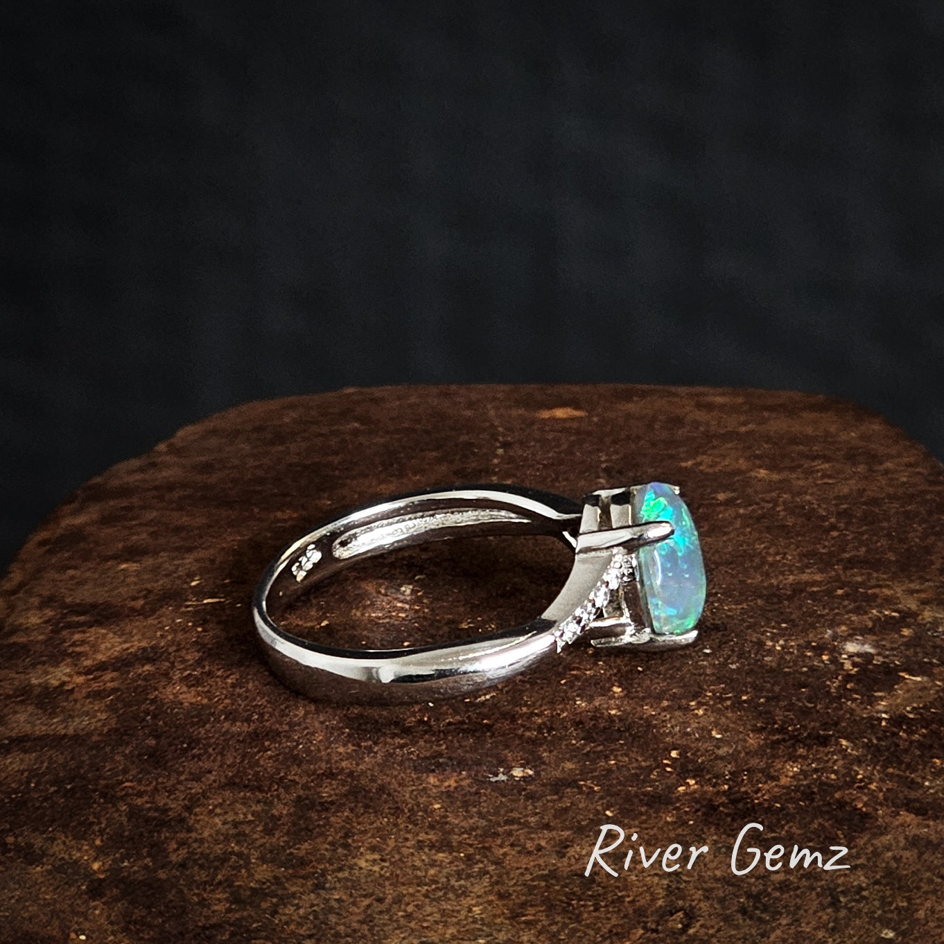 Side view of silver ring with a green gemstone on a dark surface, branded 'River Gemz'.