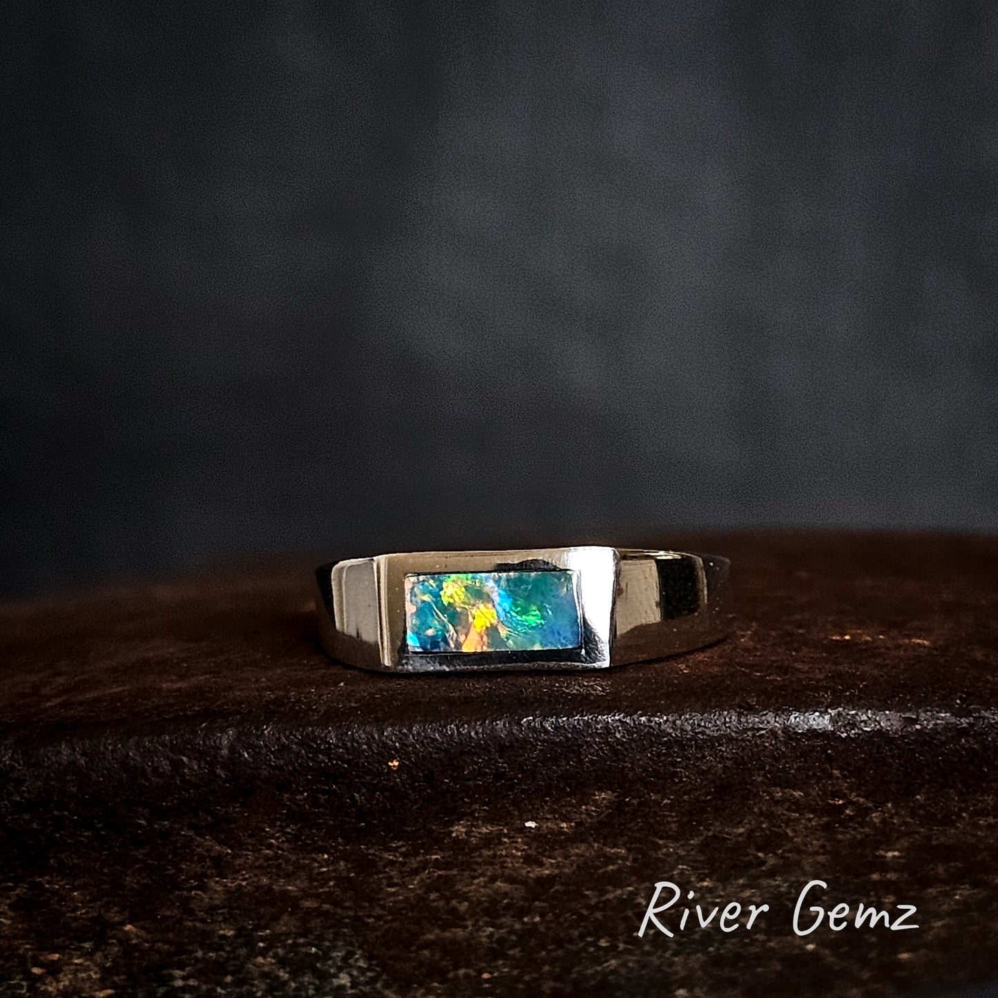 Silver ring with a colorful gemstone on a dark surface, branded 'River Gemz'.