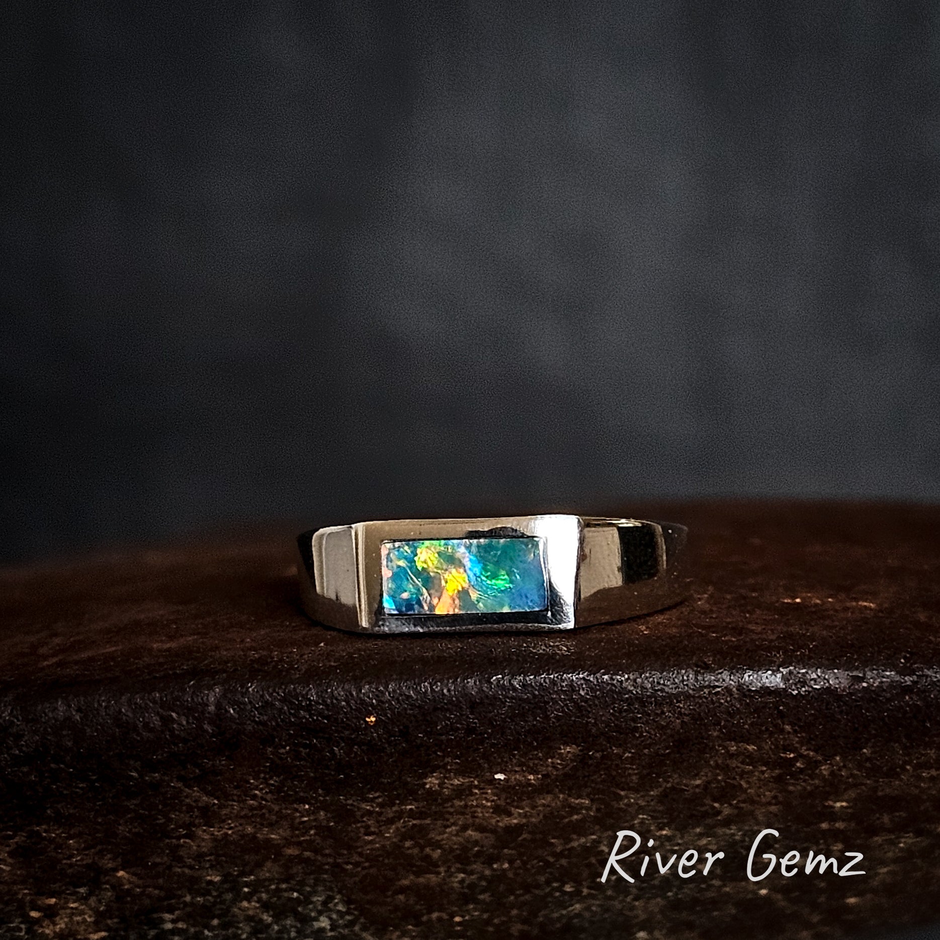 Silver ring with a colorful gemstone on a dark surface, branded 'River Gemz'.