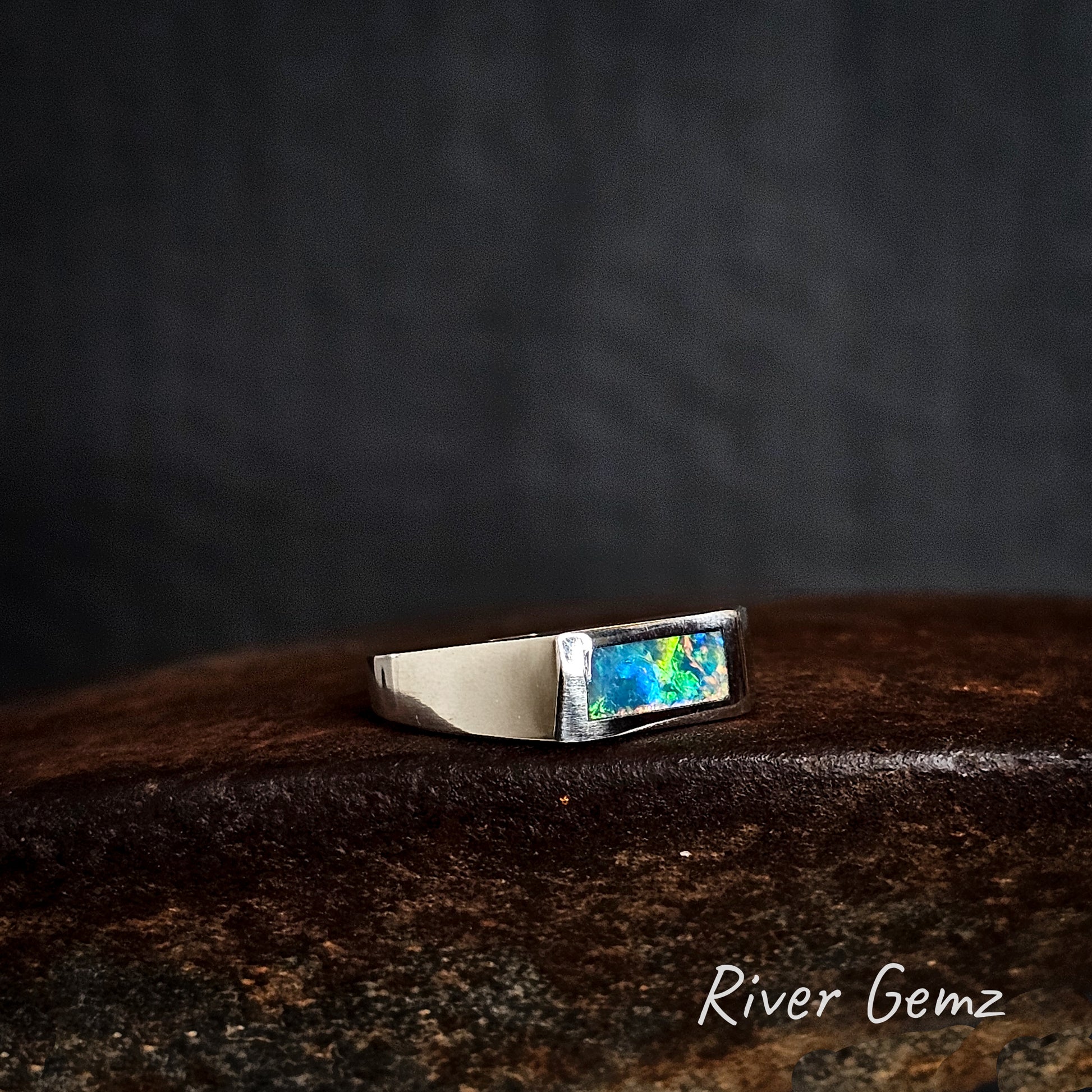 Silver ring with a colorful gemstone on a dark background, featuring the brand 'River Gemz'.