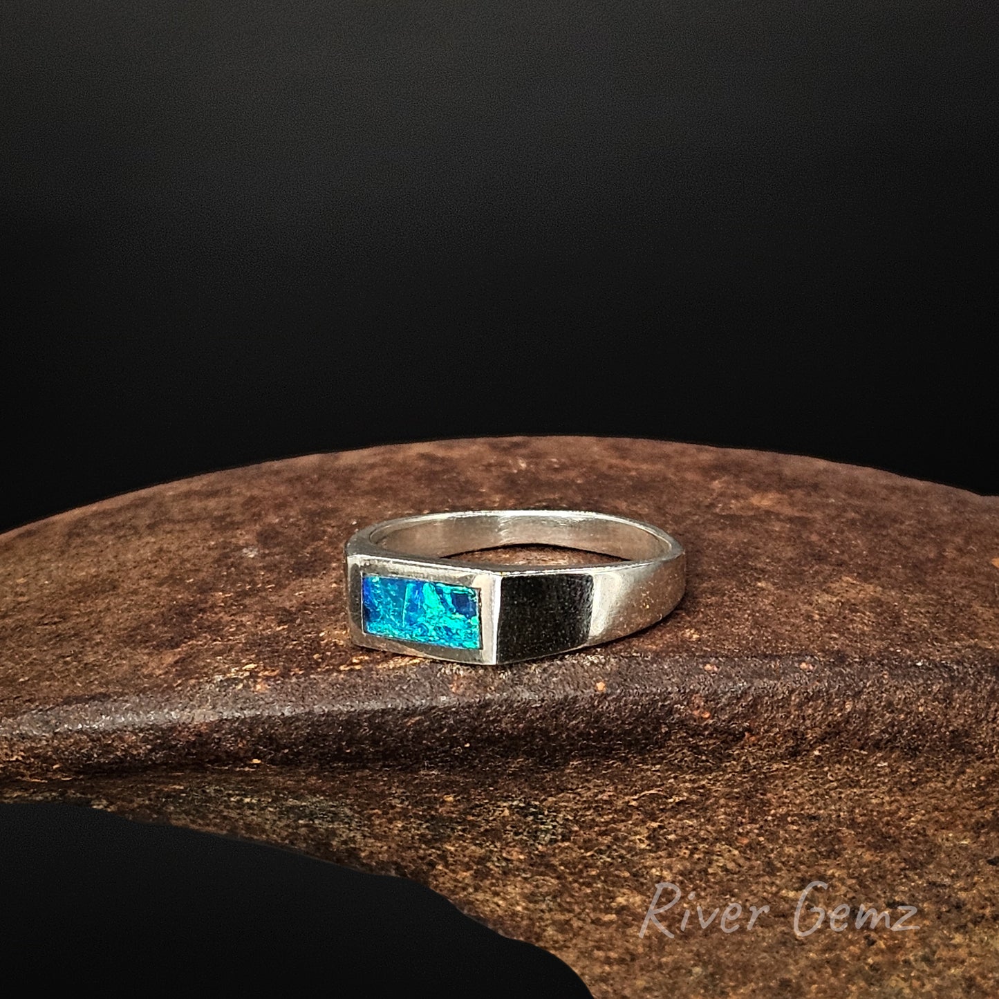 Silver ring with a blue gemstone on a wooden surface, branded 'River Gemz'.