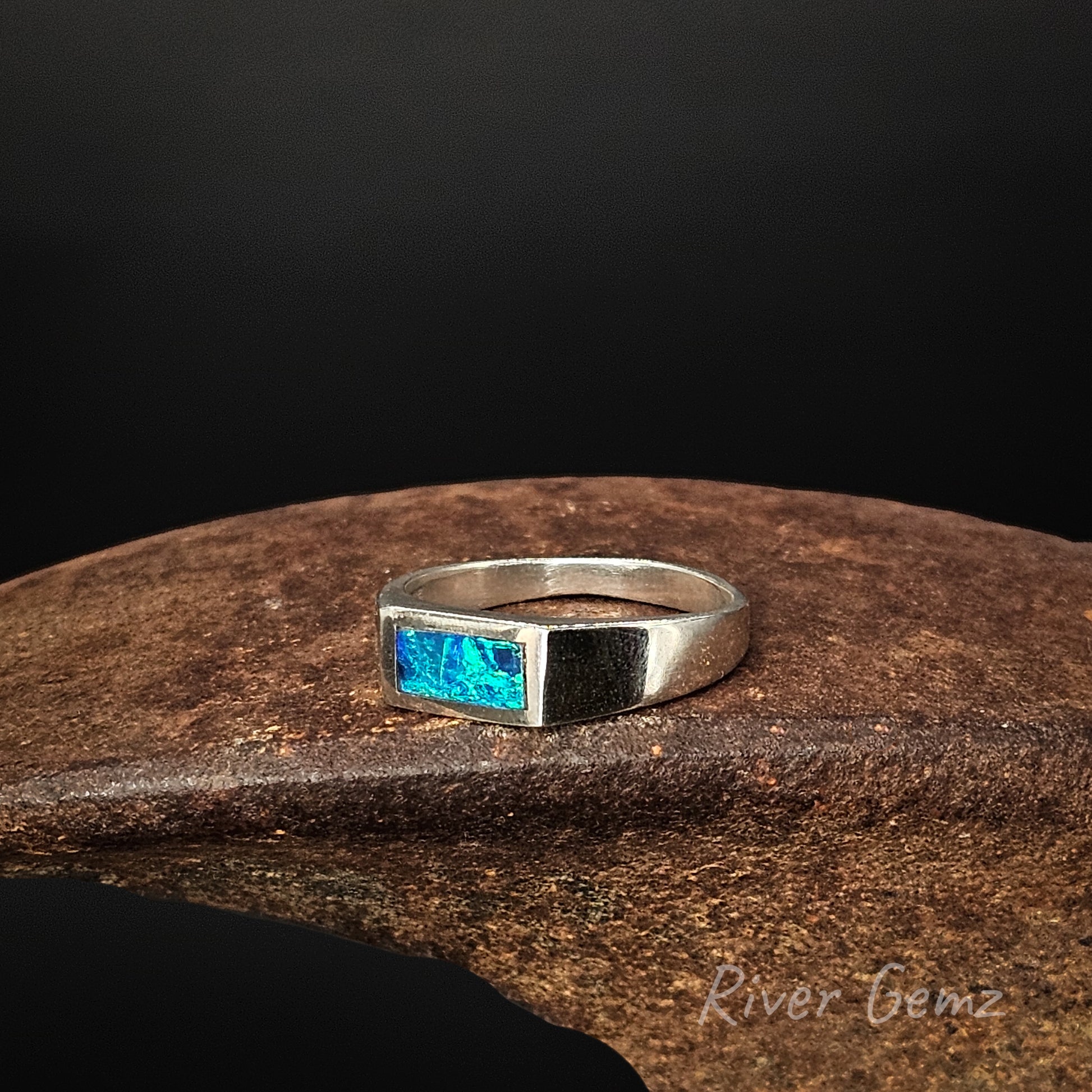 Silver ring with a blue gemstone on a wooden surface, branded 'River Gemz'.