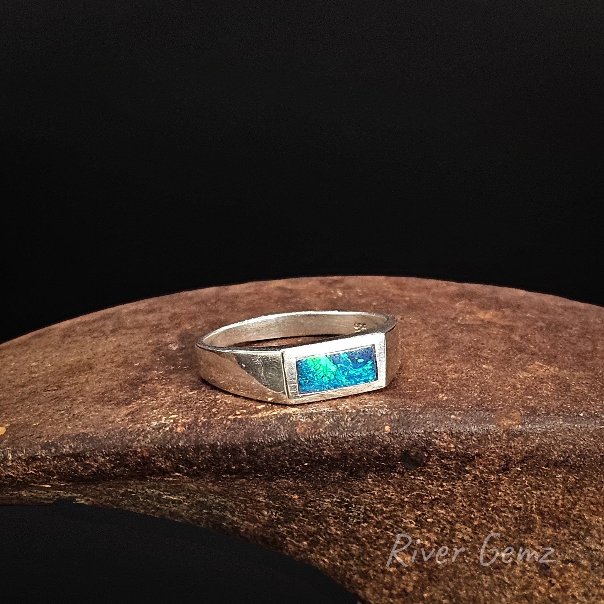 Silver ring with a blue gemstone on a dark brown surface, branded 'River Gemz'.