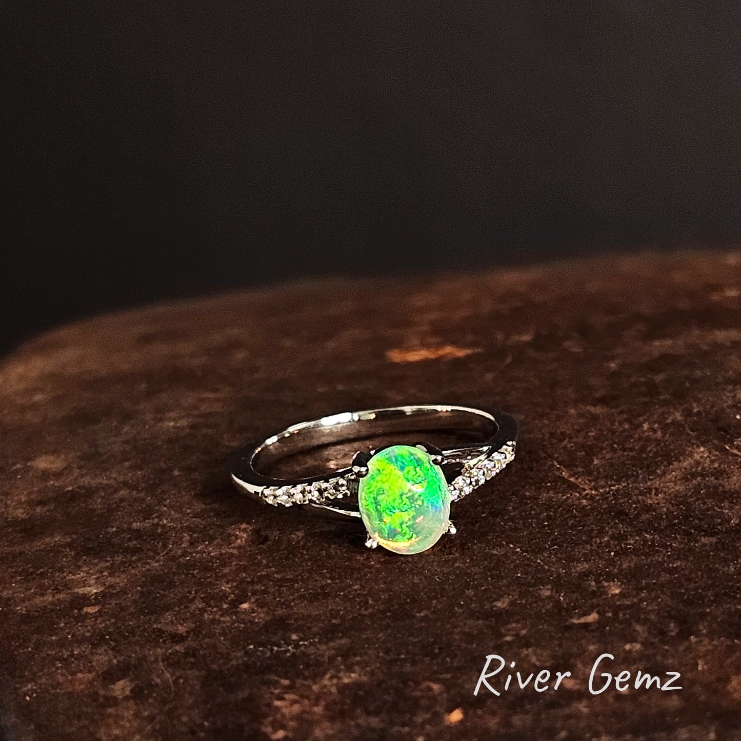 Ring with a bright green opal stone on a dark brown surface, branded 'River Gemz'.