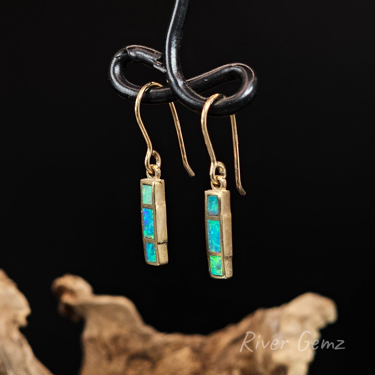 Yellow gold hook earrings with 3 rectangular shaped blue and green opals channel set. Black background and drift wood at base with 'River Gemz' watermark in the bottom right hand corner.