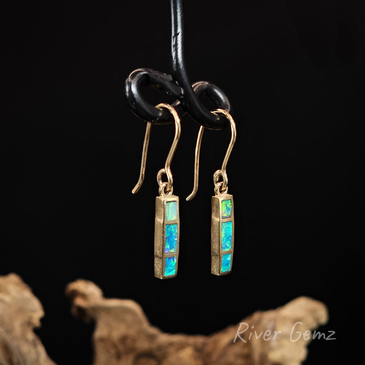 Yellow gold hook earrings with 3 rectangular shaped blue and green opals channel set. Black background and drift wood at base with 'River Gemz' watermark in the bottom right hand corner.