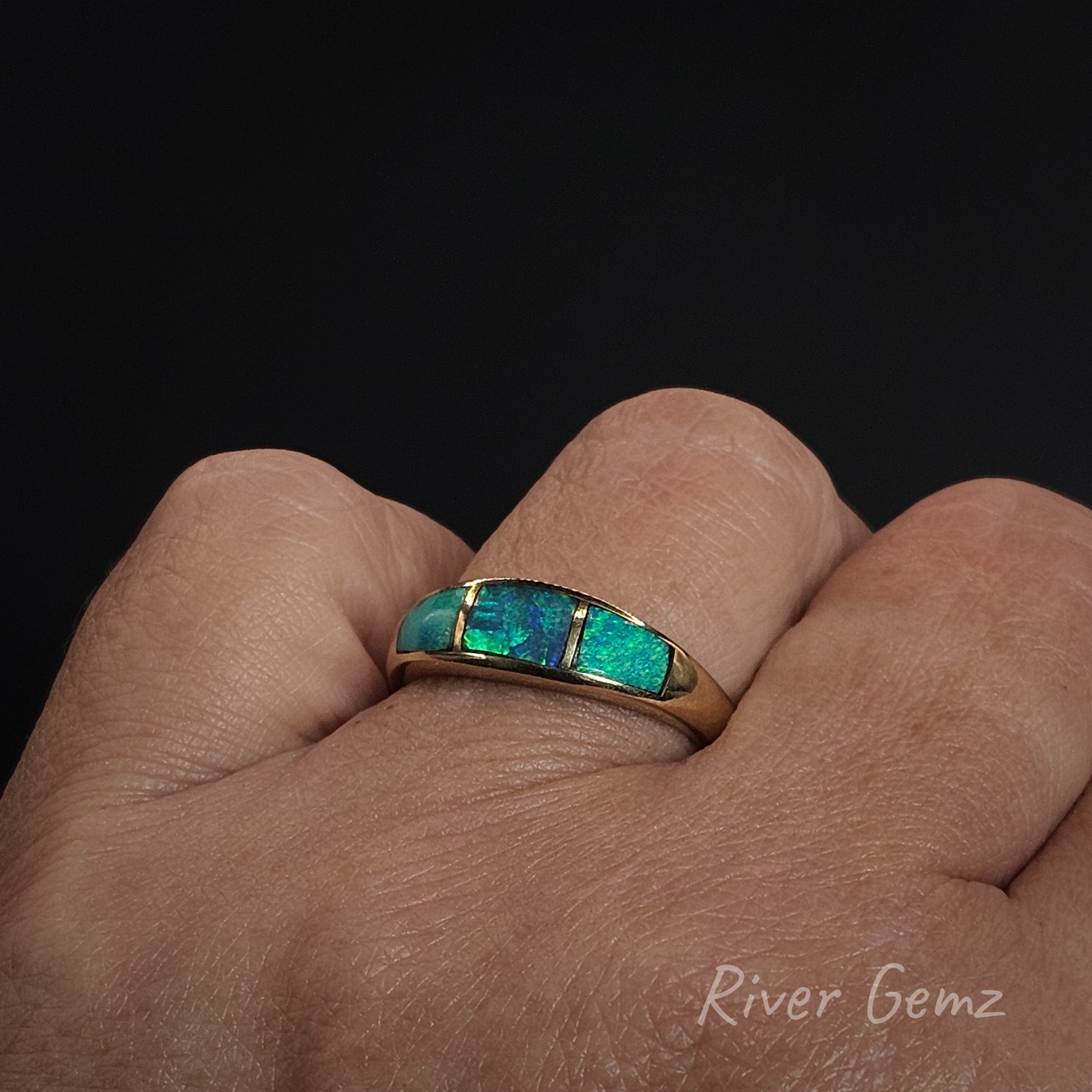 Gold ring with green gemstone on a hand against a black background, featuring 'River Gemz' branding.