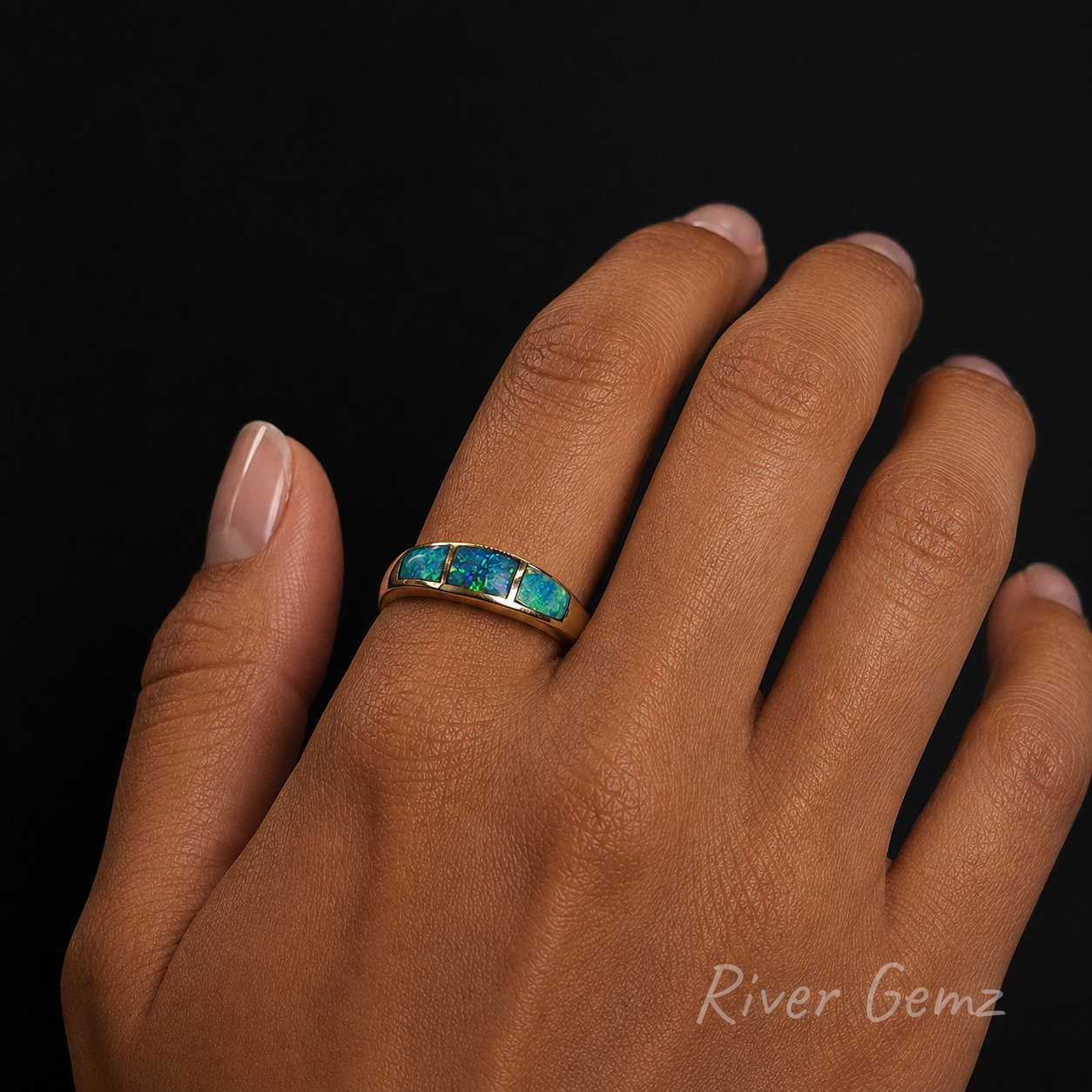 Hand wearing a gold ring with blue gemstones on a black background, branded 'River Gemz'.