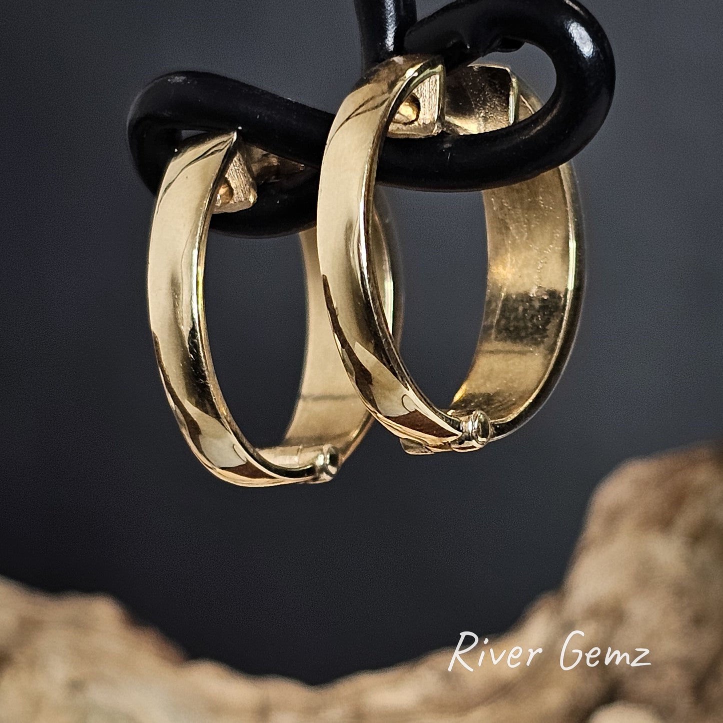 Rear of gold hoop earrings hanging on a black cord against a dark background with 'River Gemz' branding.