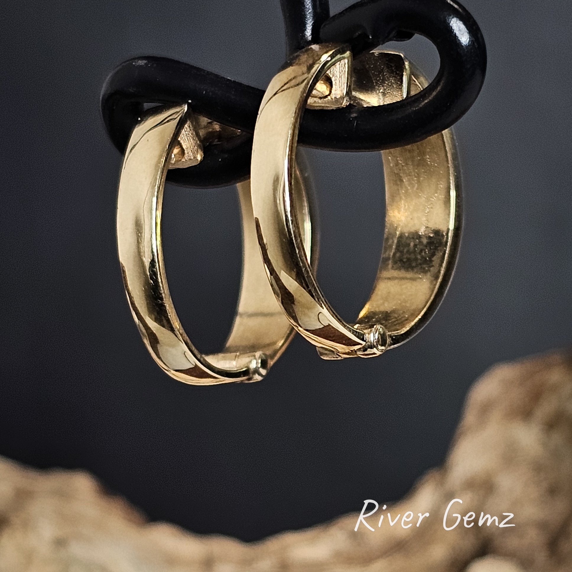 Rear of gold hoop earrings hanging on a black cord against a dark background with 'River Gemz' branding.