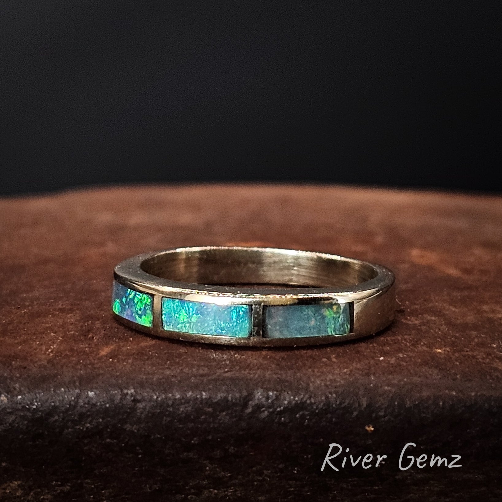 White gold ring with embedded blue-green opal stones on a dark brown surface, branded 'River Gemz'.
