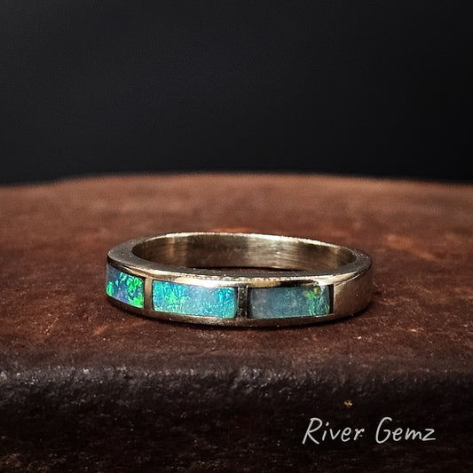White gold ring with embedded blue-green opal stones on a dark brown surface, branded 'River Gemz'.
