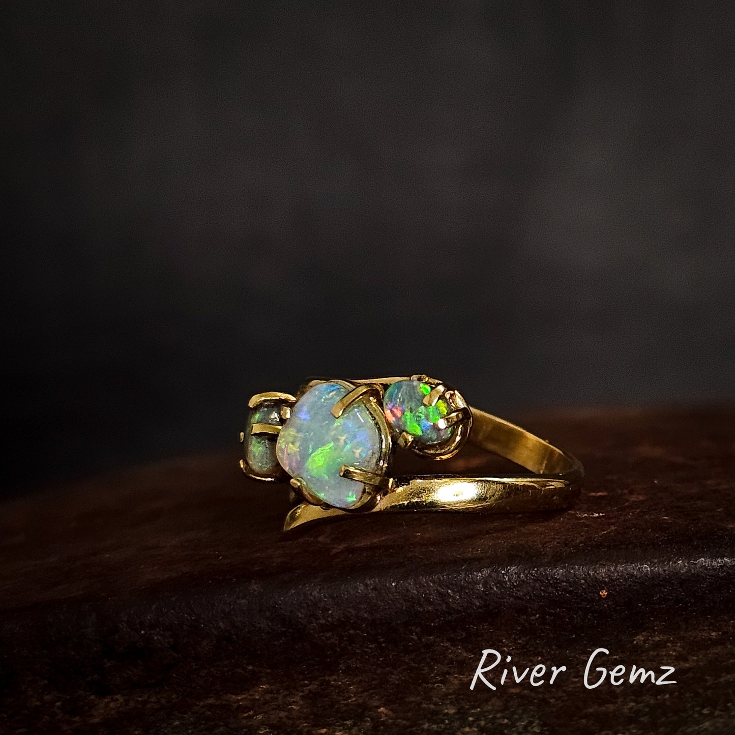 Trilogy gold ring with 3 light stones on a cast iron surface with 'River Gemz' branding.