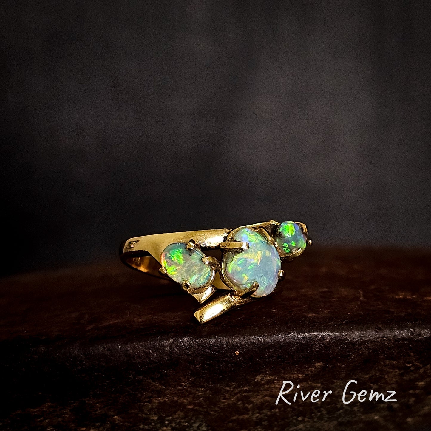 Three light opals claw set in yellow gold ring on a cast iron surface with 'River Gemz' branding.