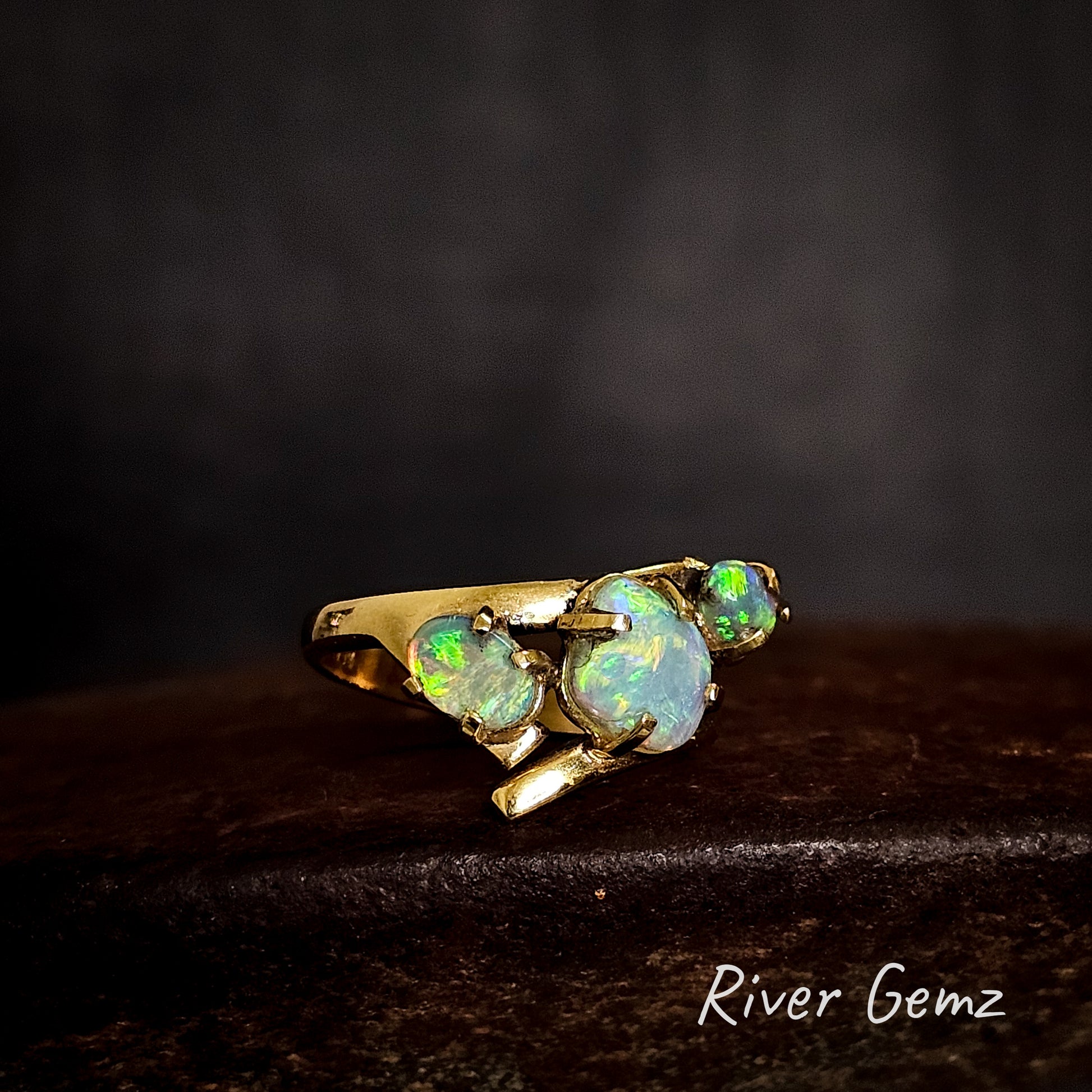 Three light opals claw set in yellow gold ring on a cast iron surface with 'River Gemz' branding.
