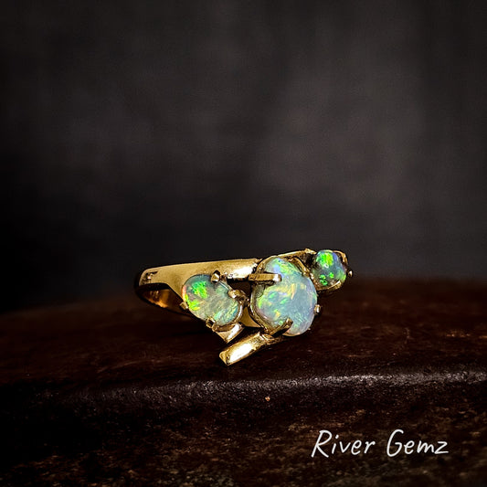 Three light opals claw set in yellow gold ring on a cast iron surface with 'River Gemz' branding.