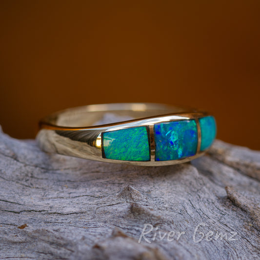 3 blue green crystal opals in yellow gold ring sitting on light grey grained wood.