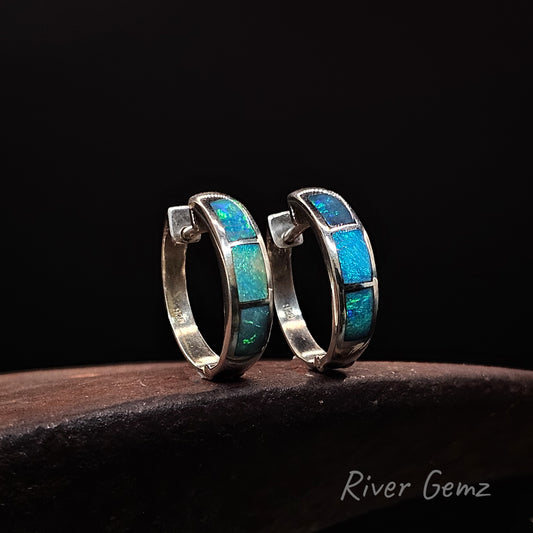 Pair of silver hoop earrings with 3 blue opals inlayed on a dark background, branded 'River Gemz'.