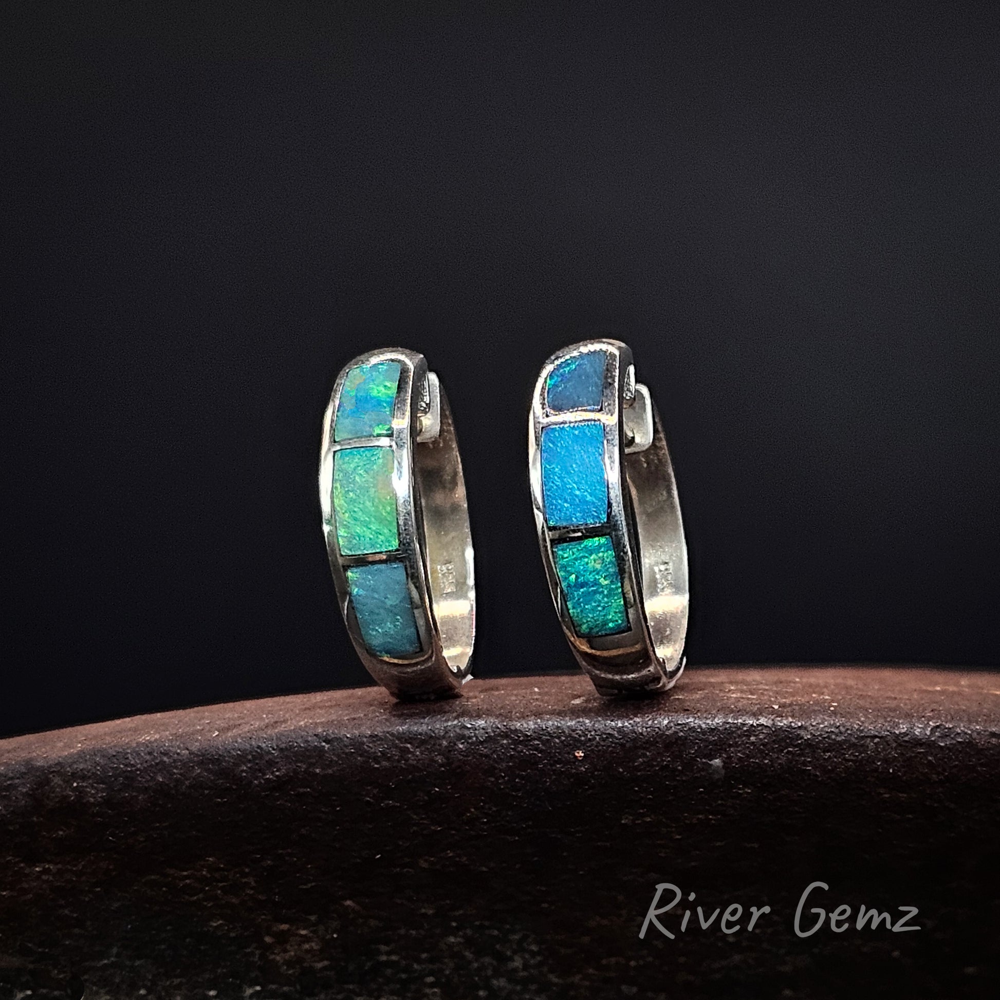 Pair of turquoise light opals inlayed in silver sleeper earrings on a dark surface with 'River Gemz' branding.
