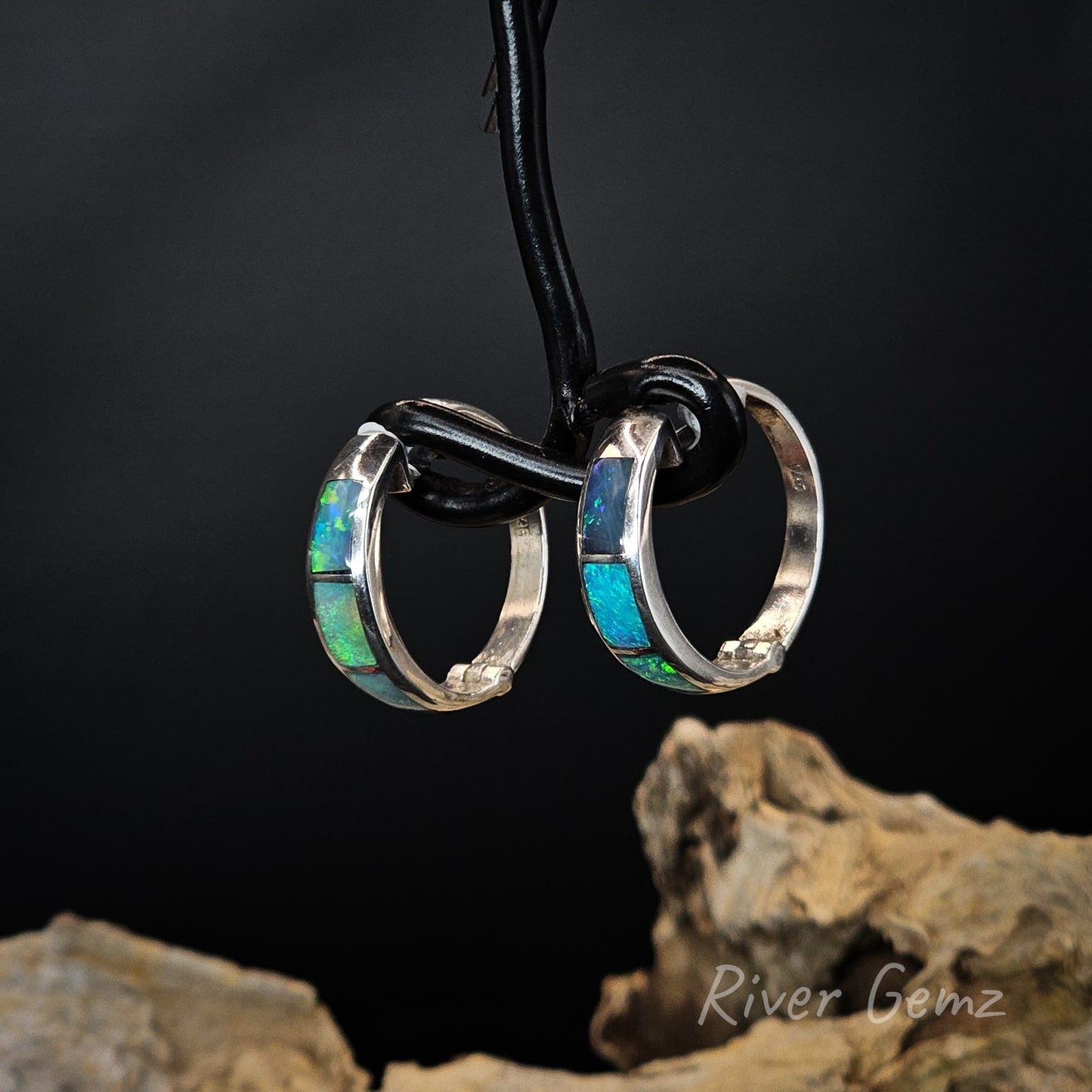Pair of huggie earrings with opal inlay on a black wire stand against a dark background.