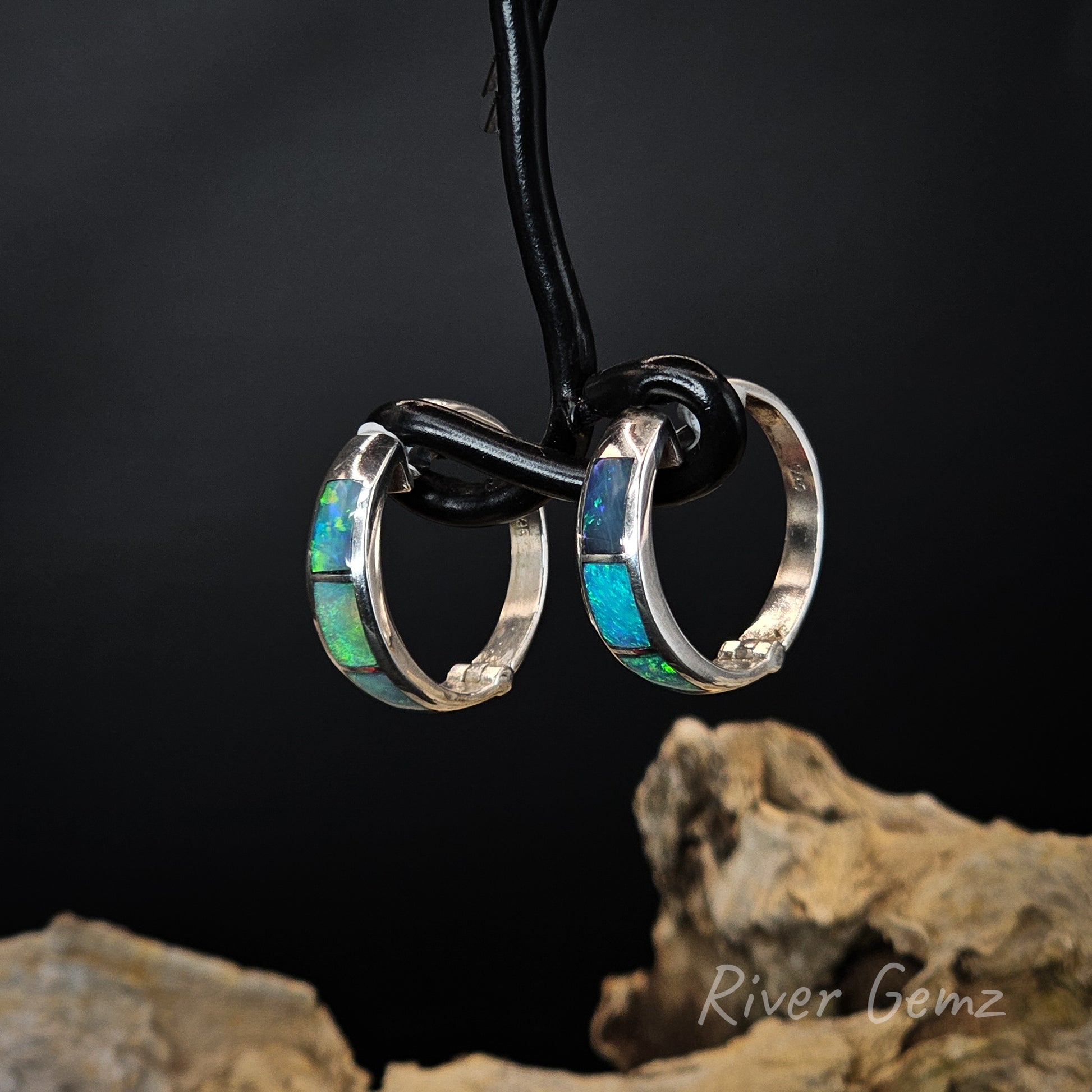 Pair of huggie earrings with opal inlay on a black wire stand against a dark background.