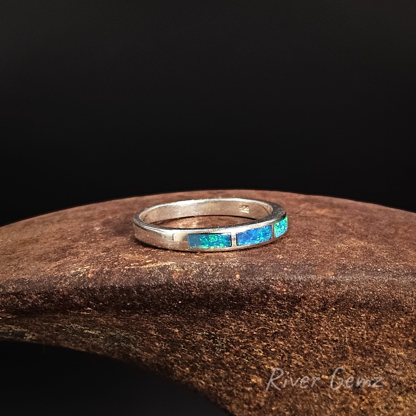Silver ring with blue stones on a dark brown surface, branded 'River Gemz'.