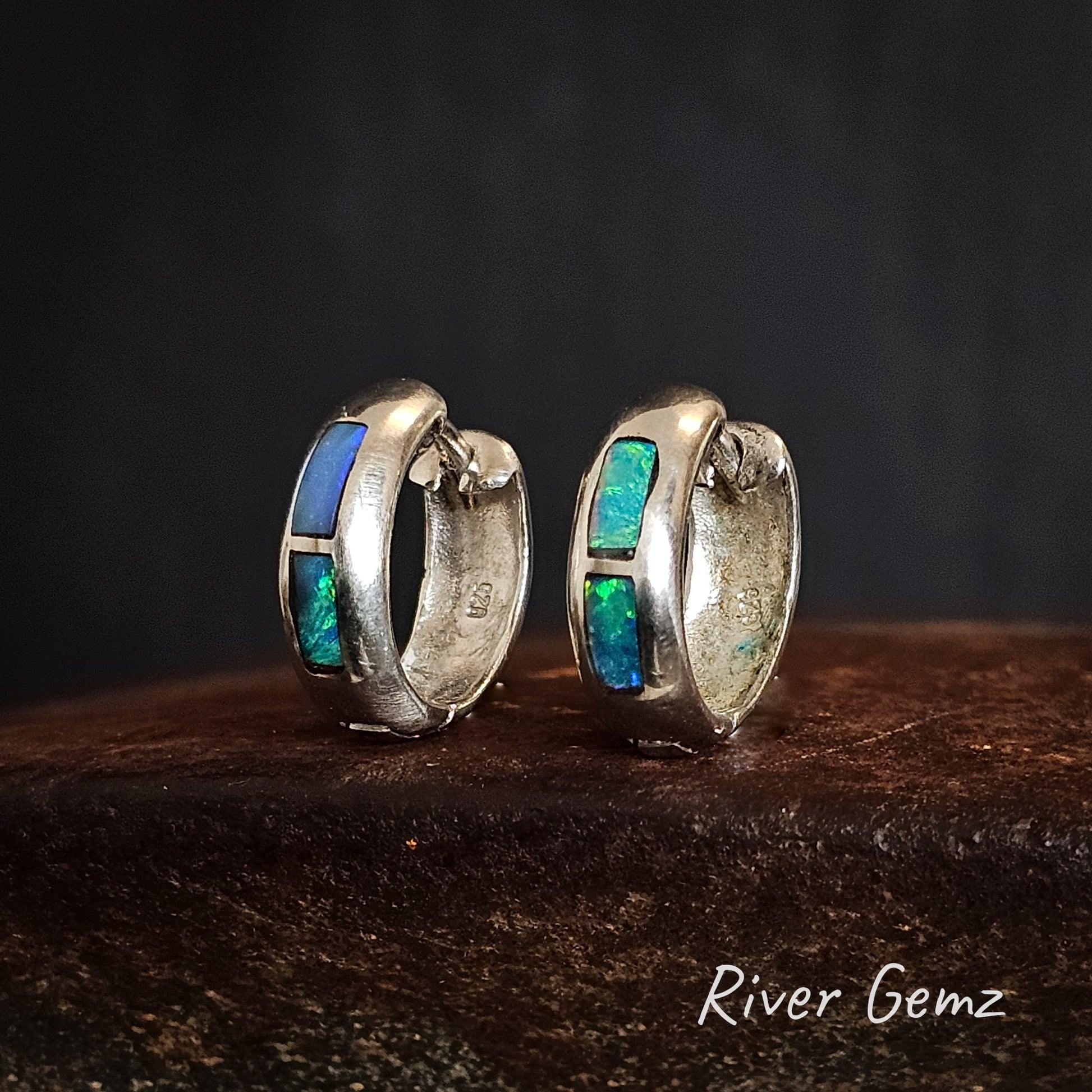 Pair of silver hoop earrings with two blue and green opals inlayed on a dark surface, branded 'River Gemz'.