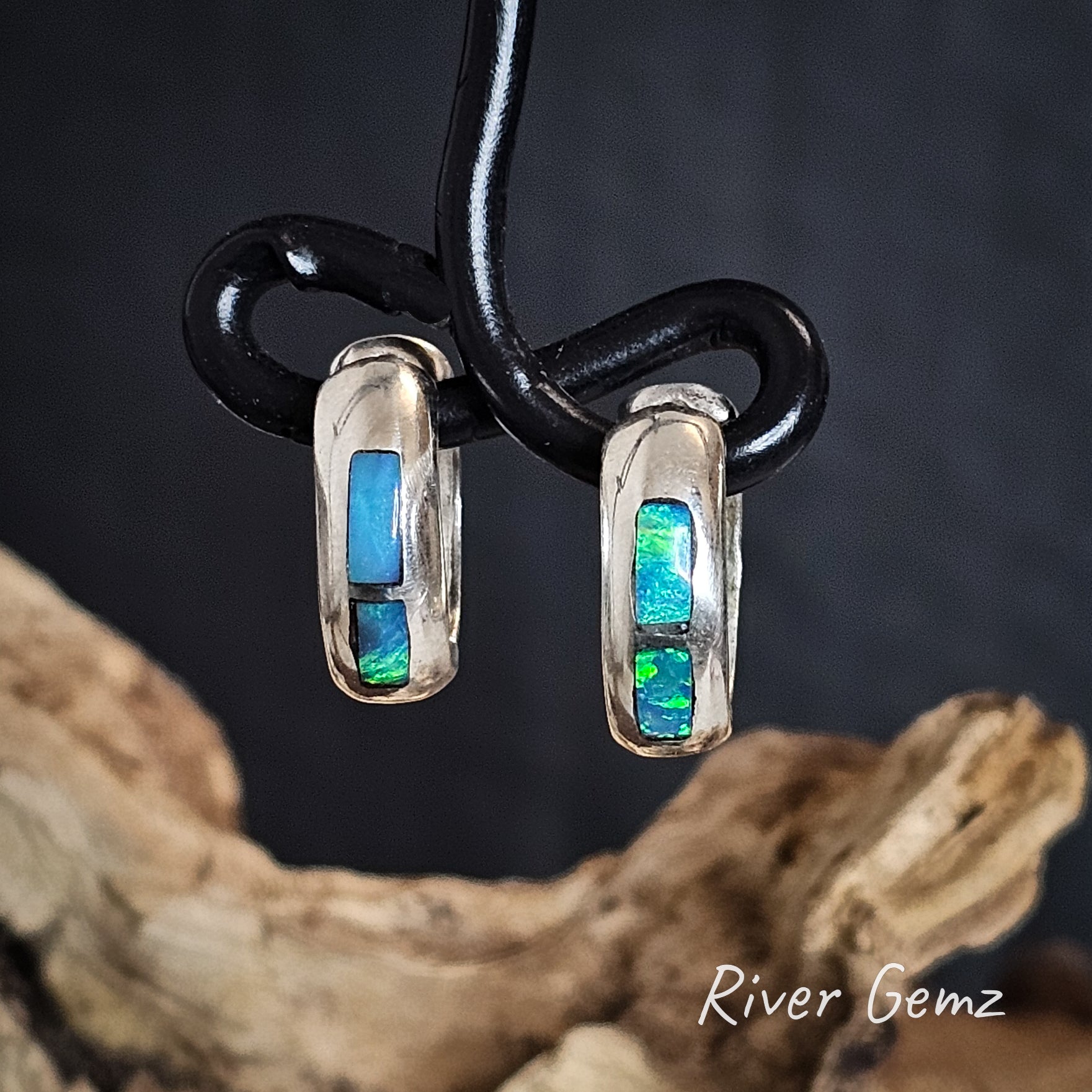 Pair of silver earrings with blue and green gemstones hanging on a black wire, set against a dark background with 'River Gemz' branding.
