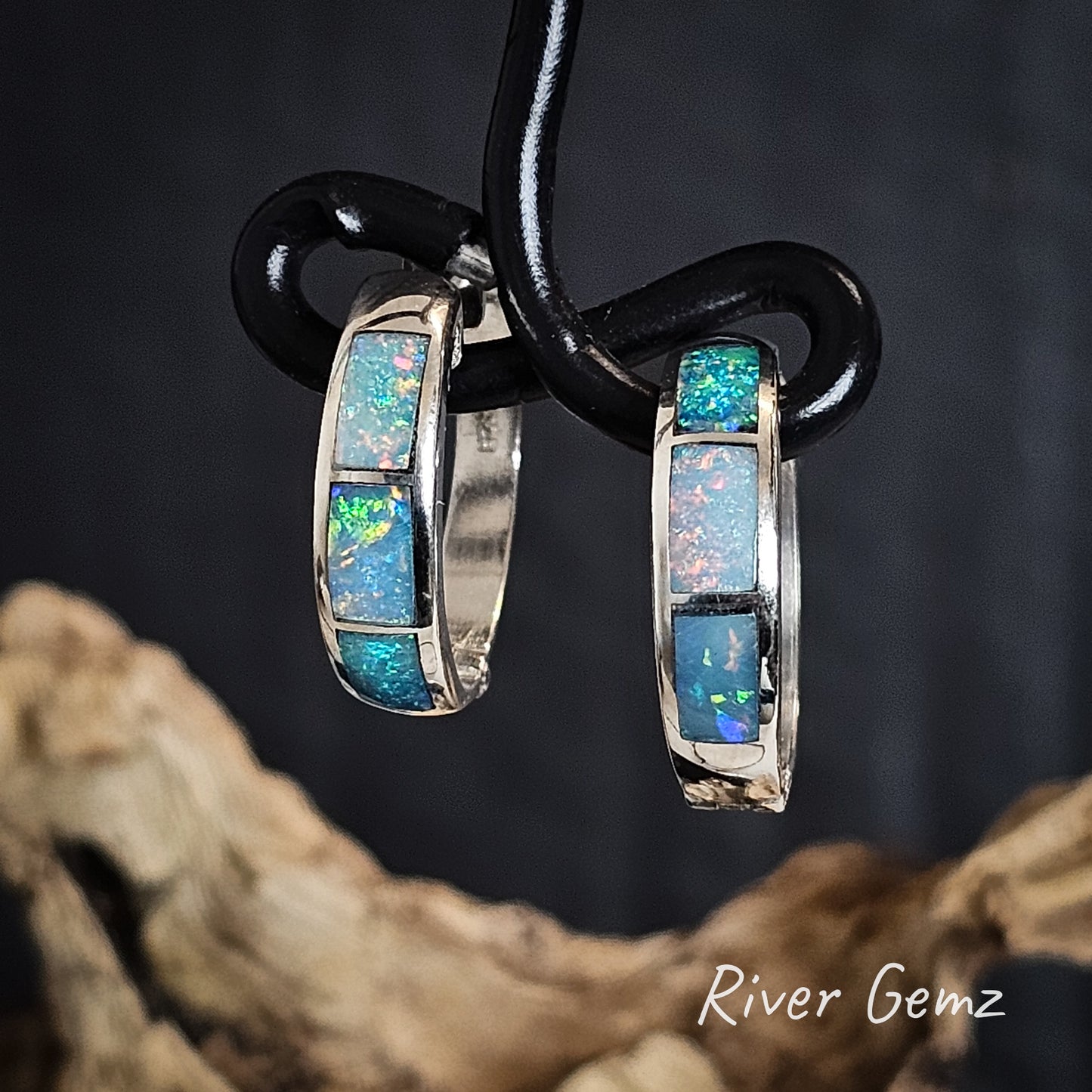 Pair of hoop earrings with colourful opals on a black wire holder, set against a dark background with 'River Gemz' branding.