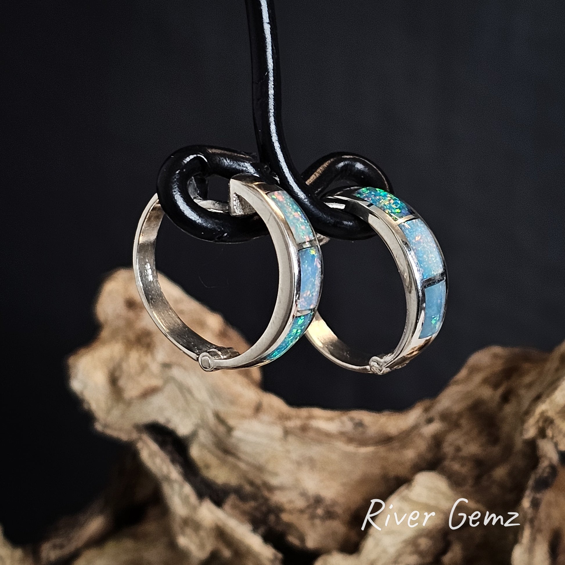 Side view of silver hoop earrings with three opals on a black wire holder, displayed on a natural wood surface with 'River Gemz' branding.