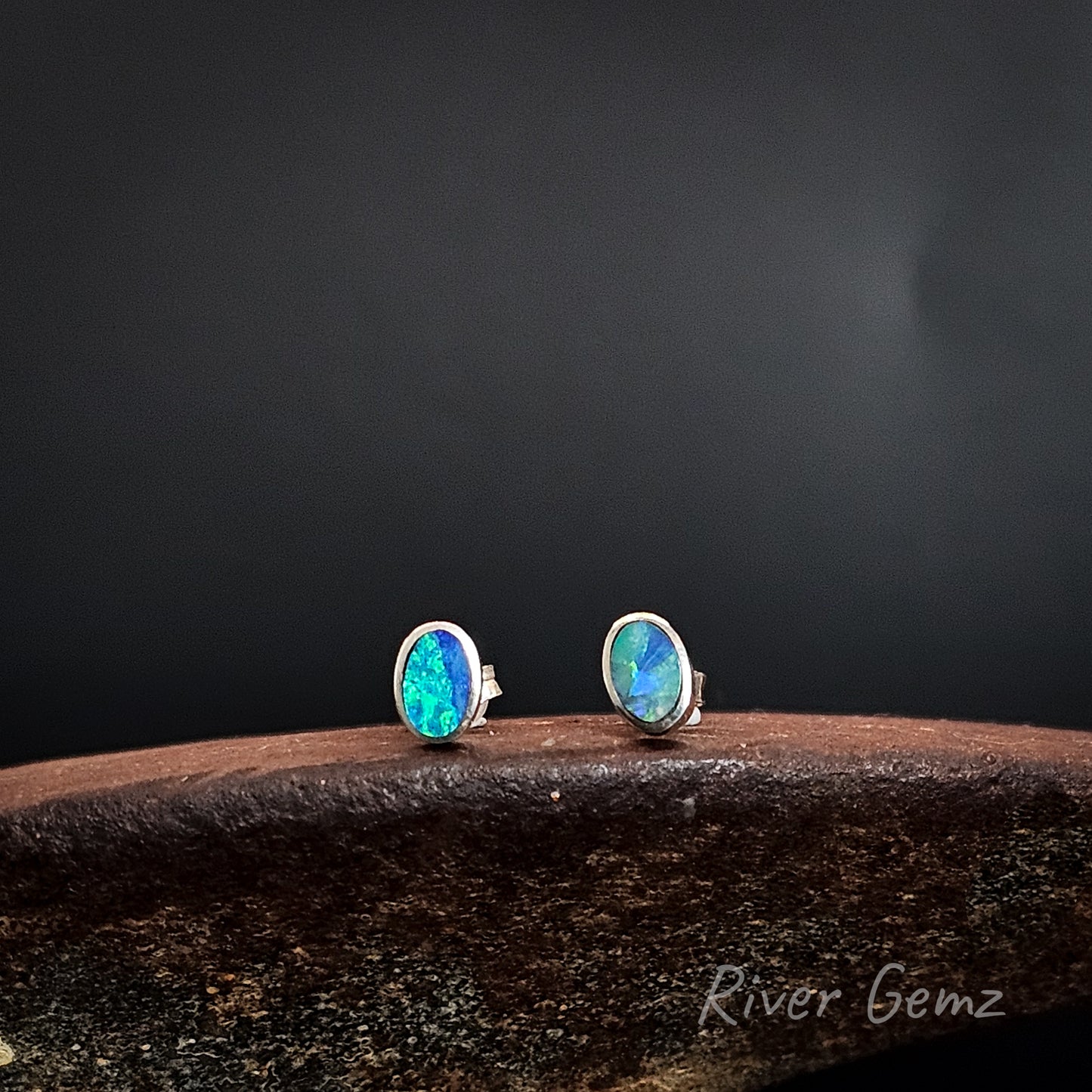 Pair of oval shaped blue and green opals set in silver stud earrings on a dark background with 'River Gemz' branding.