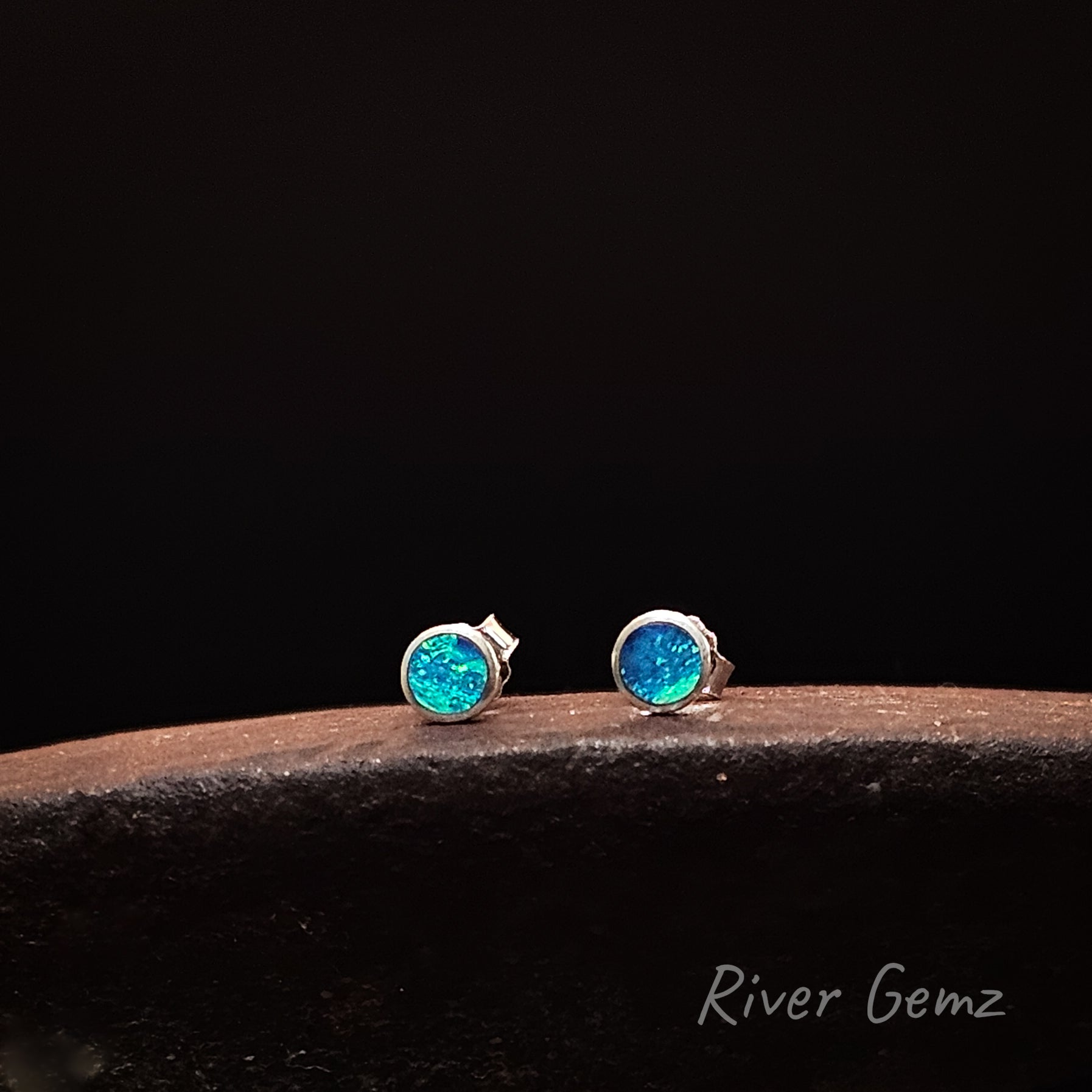 Pair of blue and green round opal stud silver earrings on a dark surface with 'River Gemz' branding.