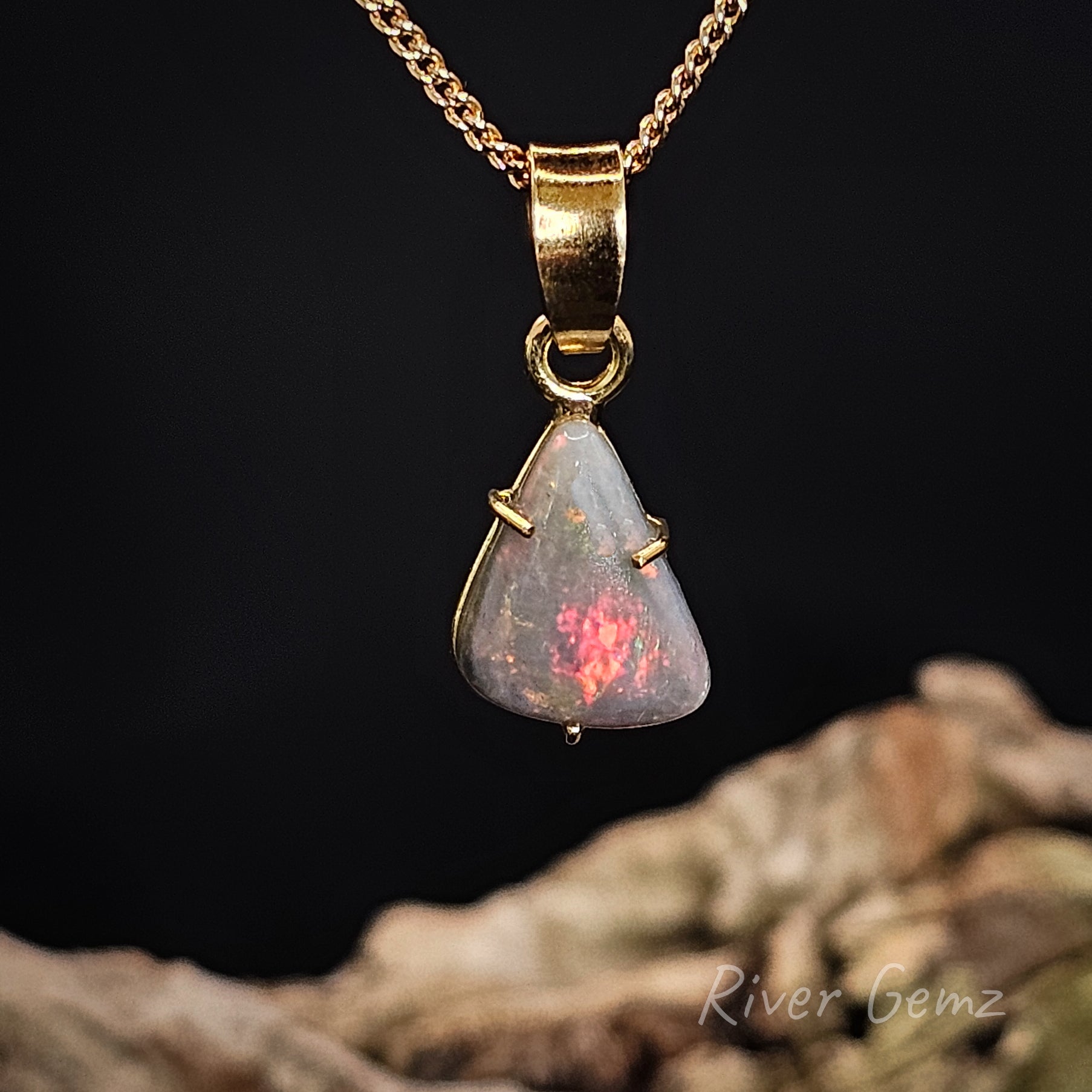 Opal with fire red on a dark opal on a gold chain with a dark background with 'River Gemz' branding.